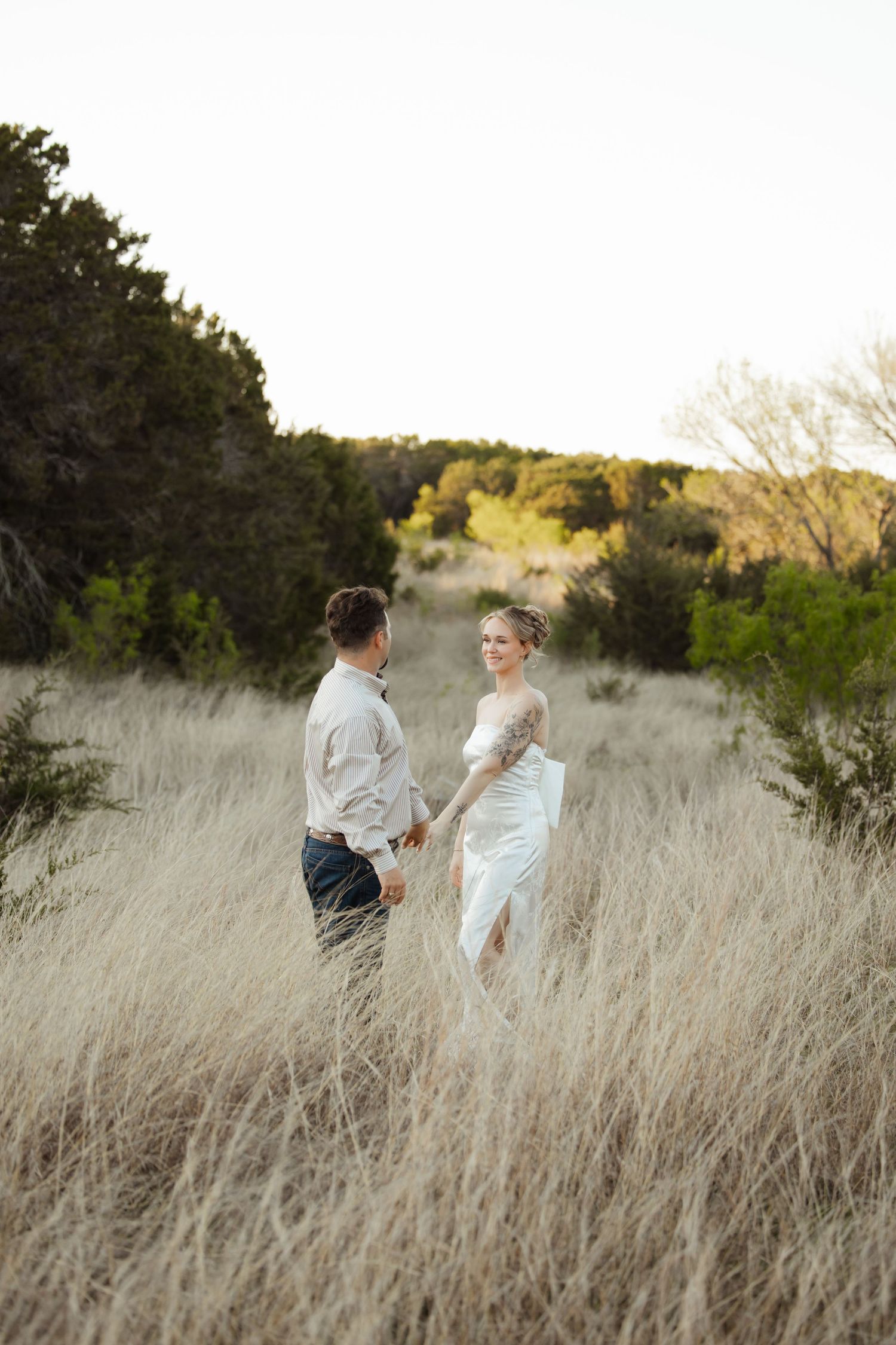 Bride and groom hold hands in golden meadow with trees at sunset during outdoor wedding photography session.