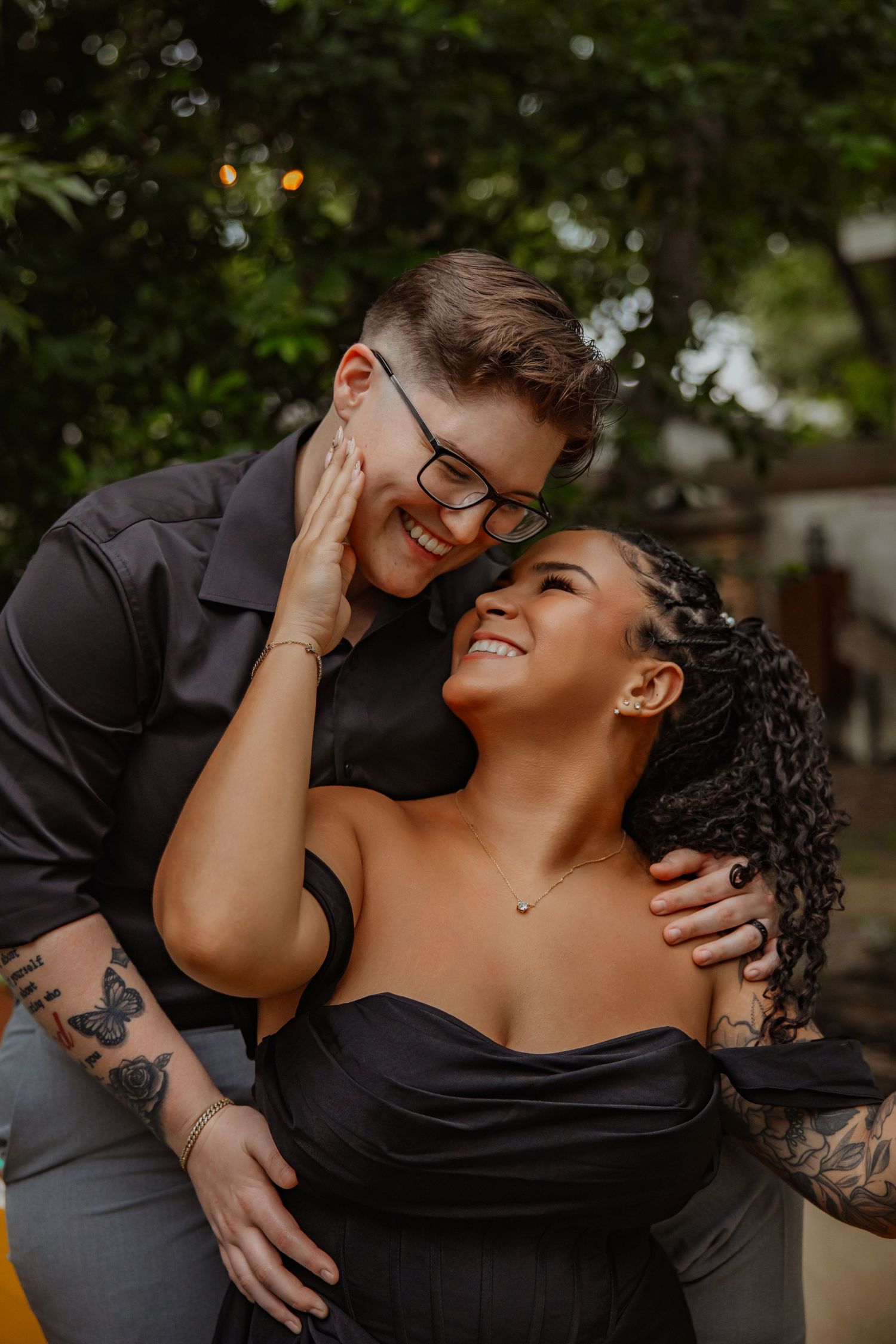 Two people in black outfits share a tender moment outdoors, smiling and embracing among lush green trees.