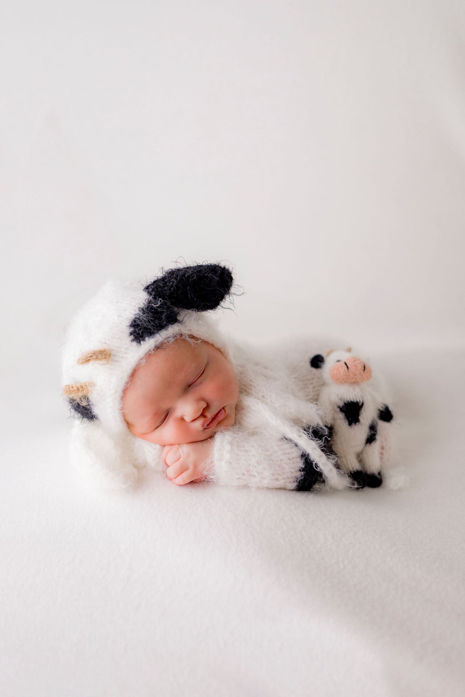 Newborn baby in black and white cow knit outfit sleeping with matching stuffed cow toy.