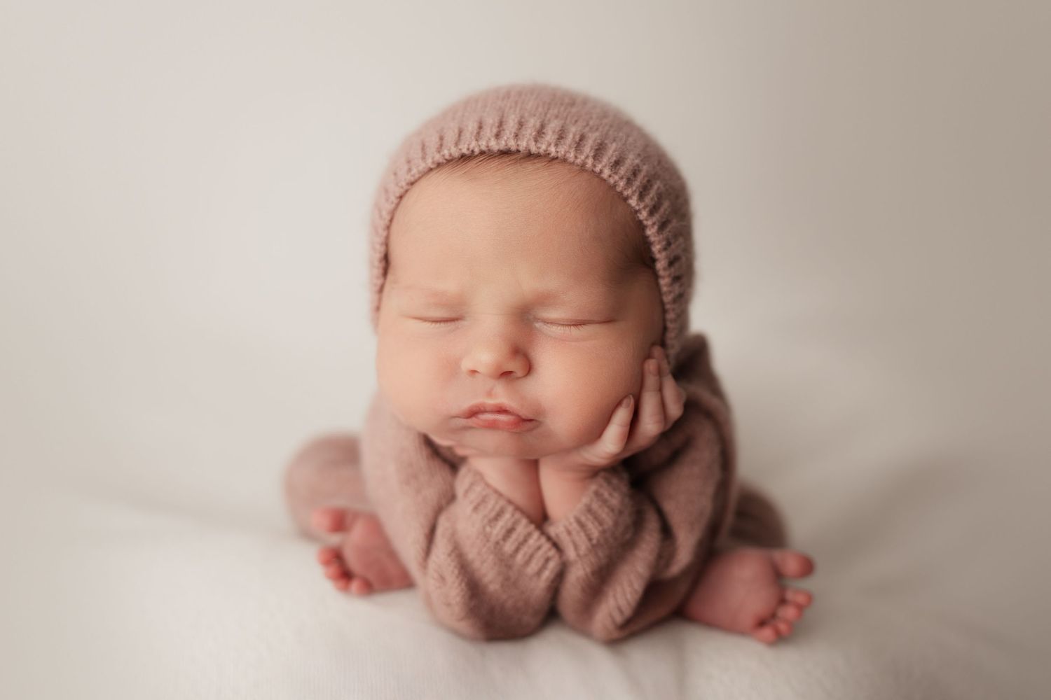 Sleeping newborn baby in dusty pink knit bonnet and romper posed with hands on cheeks.