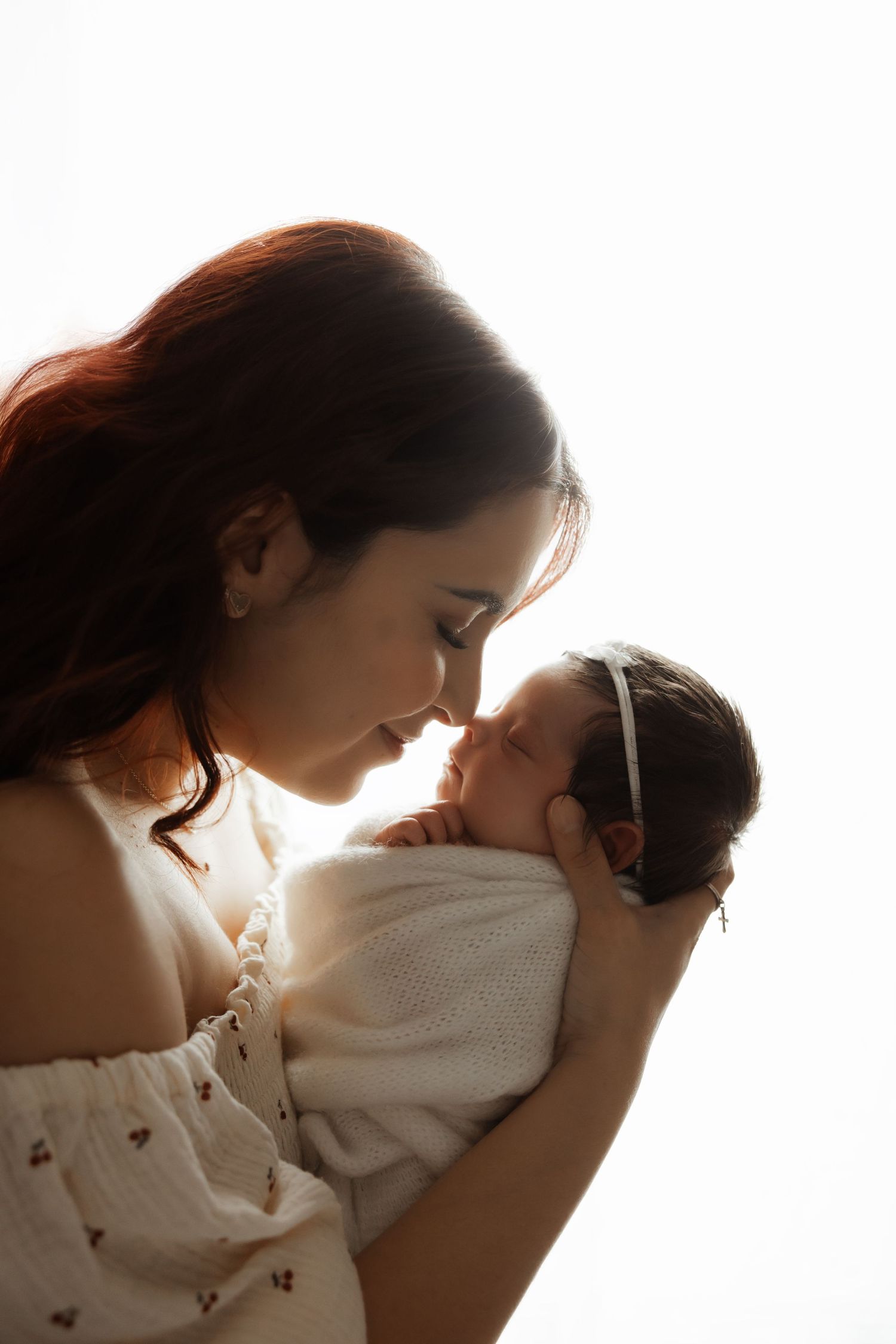 Mother in white dress tenderly touches noses with her sleeping swaddled newborn baby in a beautiful silhouette.