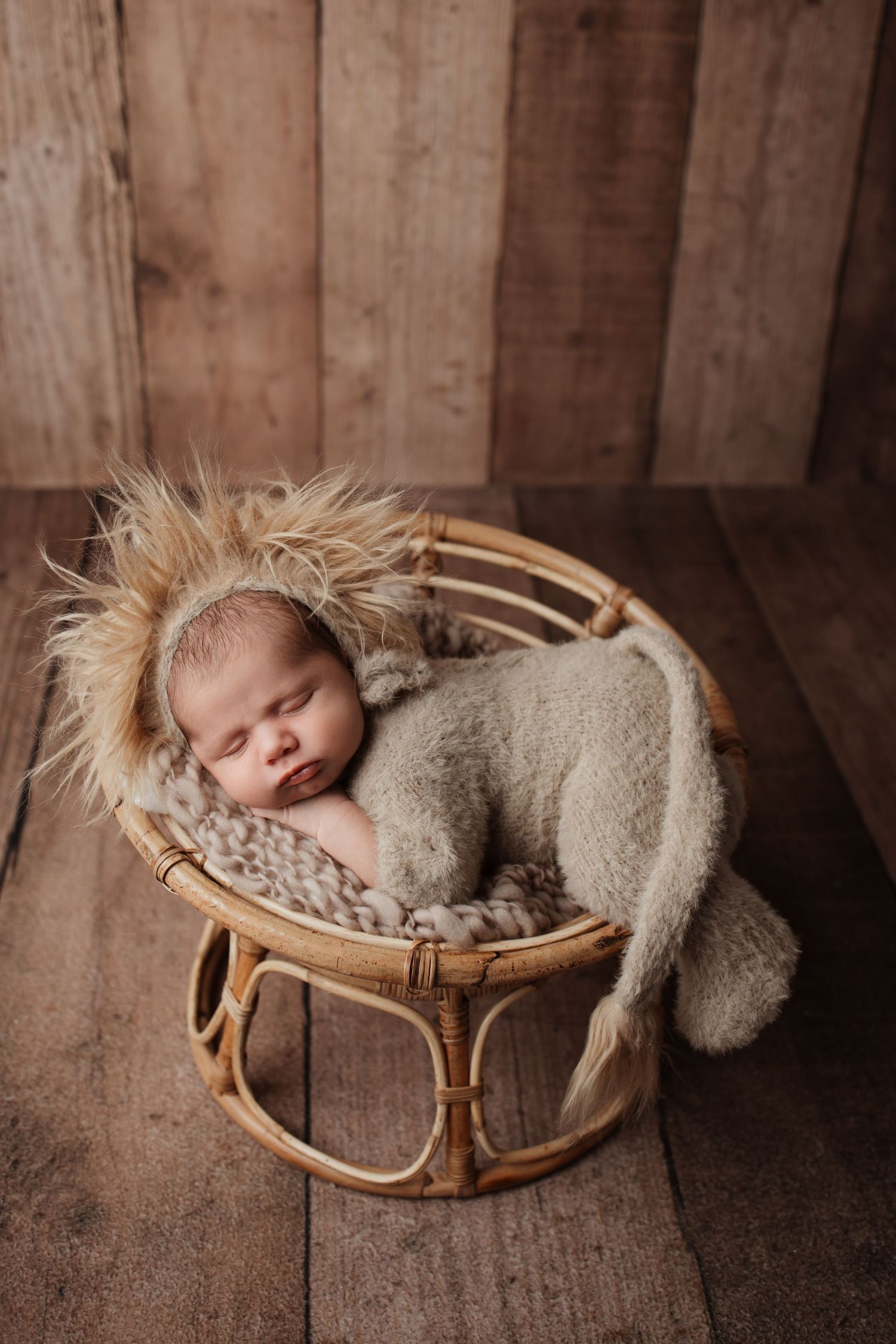 Adorable sleeping newborn wearing fluffy lion bonnet rests in a round rattan basket on wood floor.