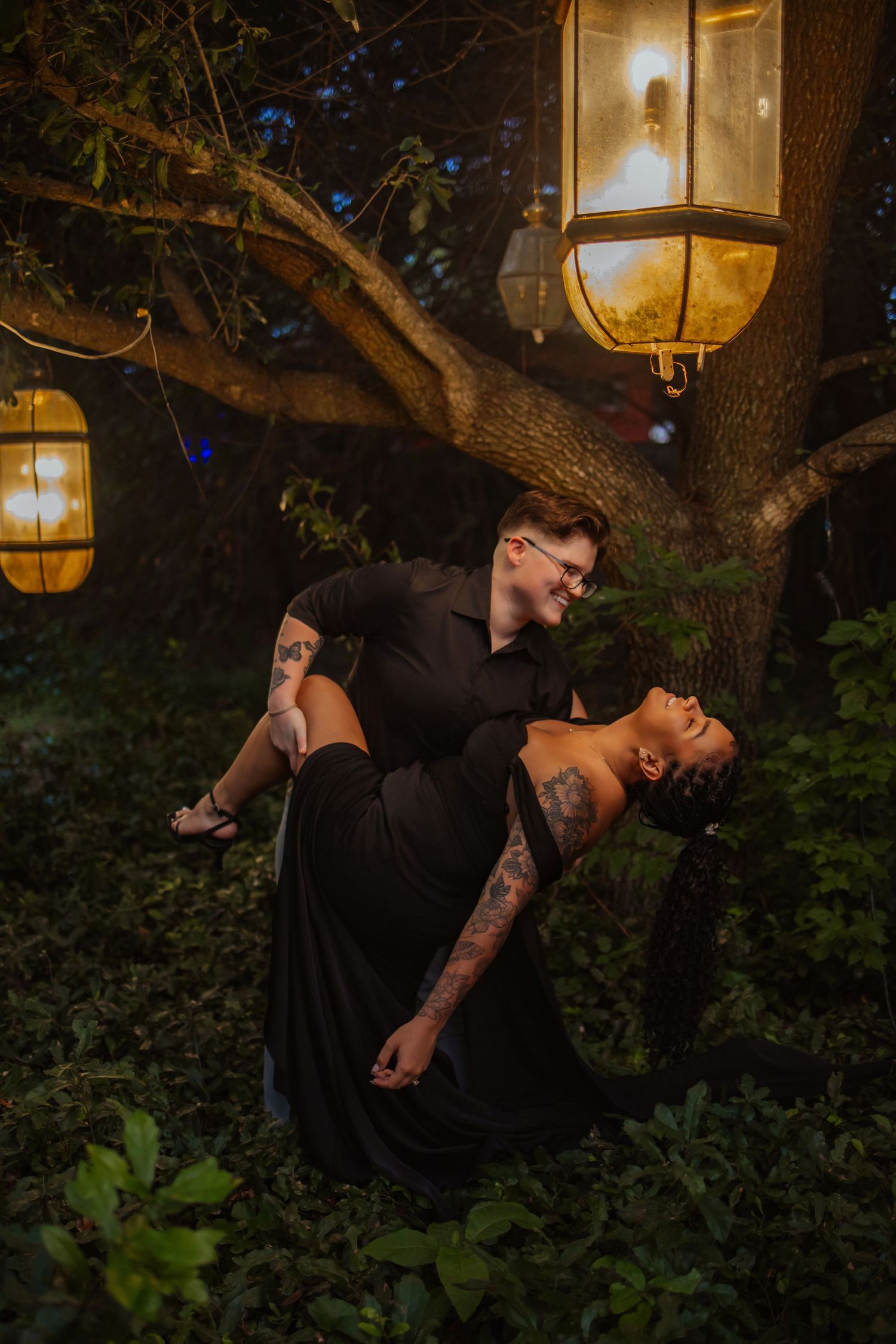 Couple in black outfits share a romantic dip pose under glowing lanterns in a lush garden at night.