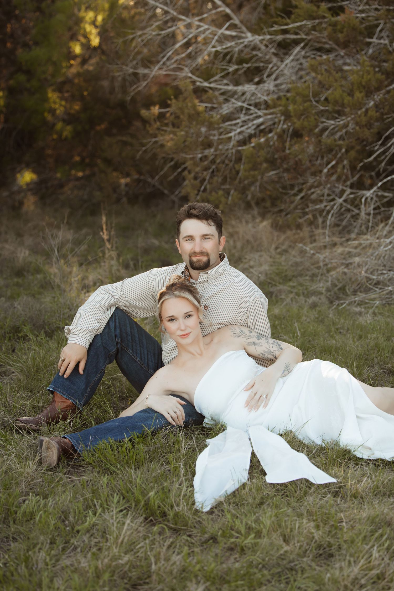 Couple sitting in grassy field, woman in white dress leaning against man in beige shirt and jeans, outdoor portrait.