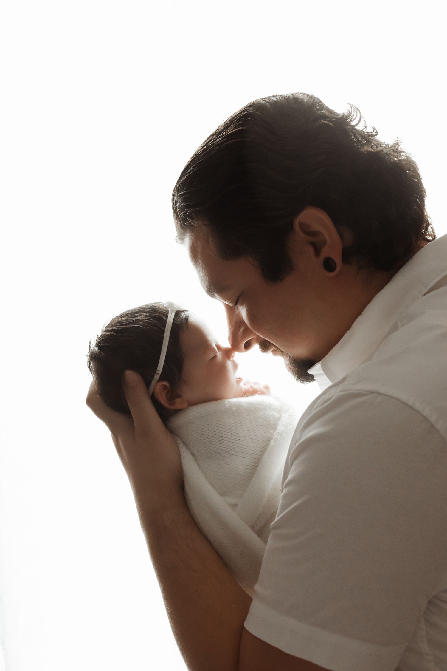Father in white shirt gently holds swaddled newborn baby, sharing an intimate nose-to-nose moment in silhouette.