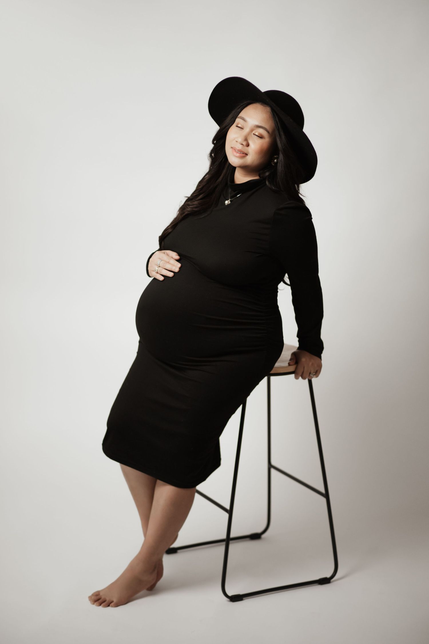 Pregnant woman in black dress and hat leaning on a stool against white background.