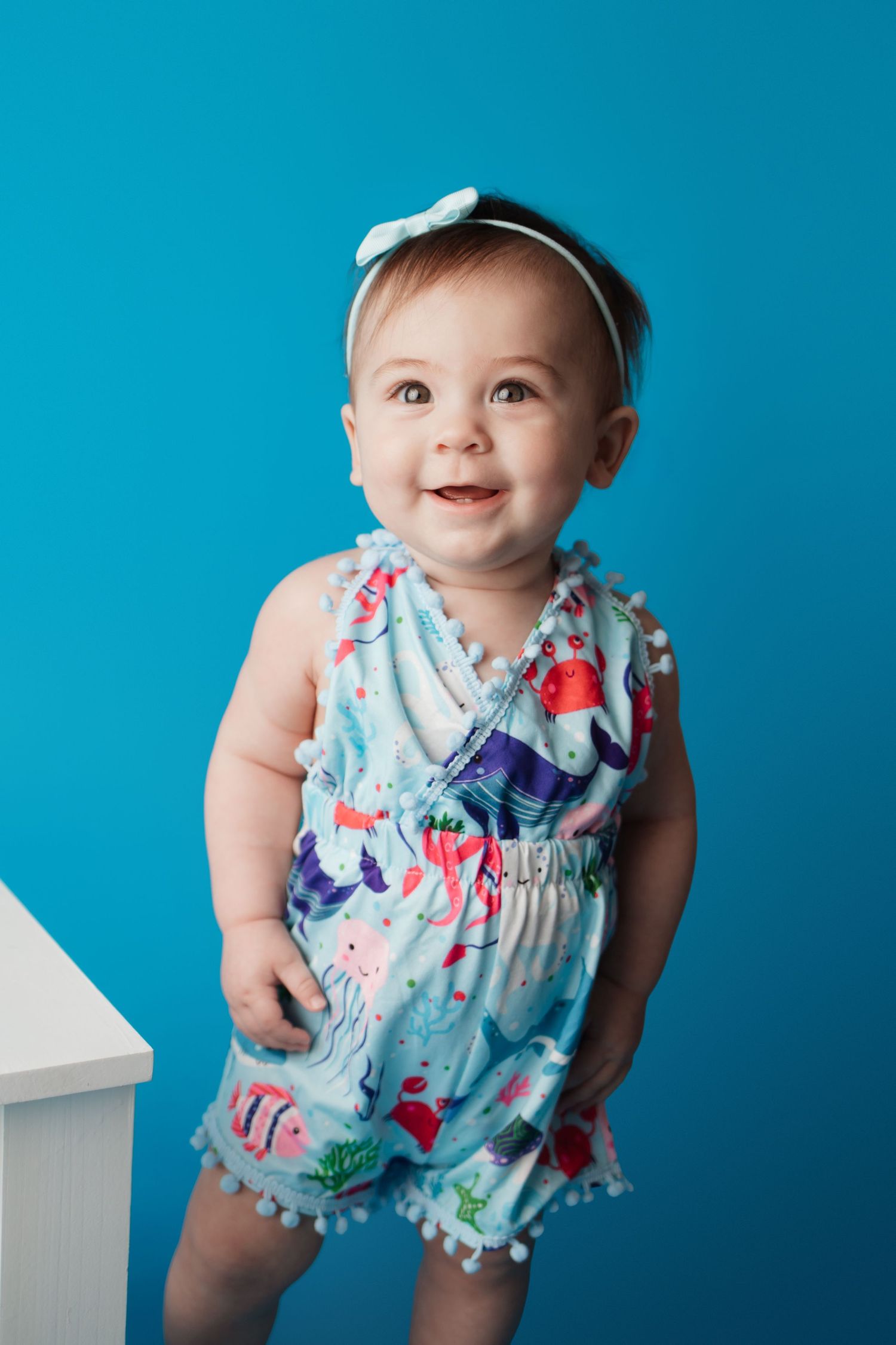 Happy baby girl in colorful ocean-themed romper with bow headband standing against blue backdrop.