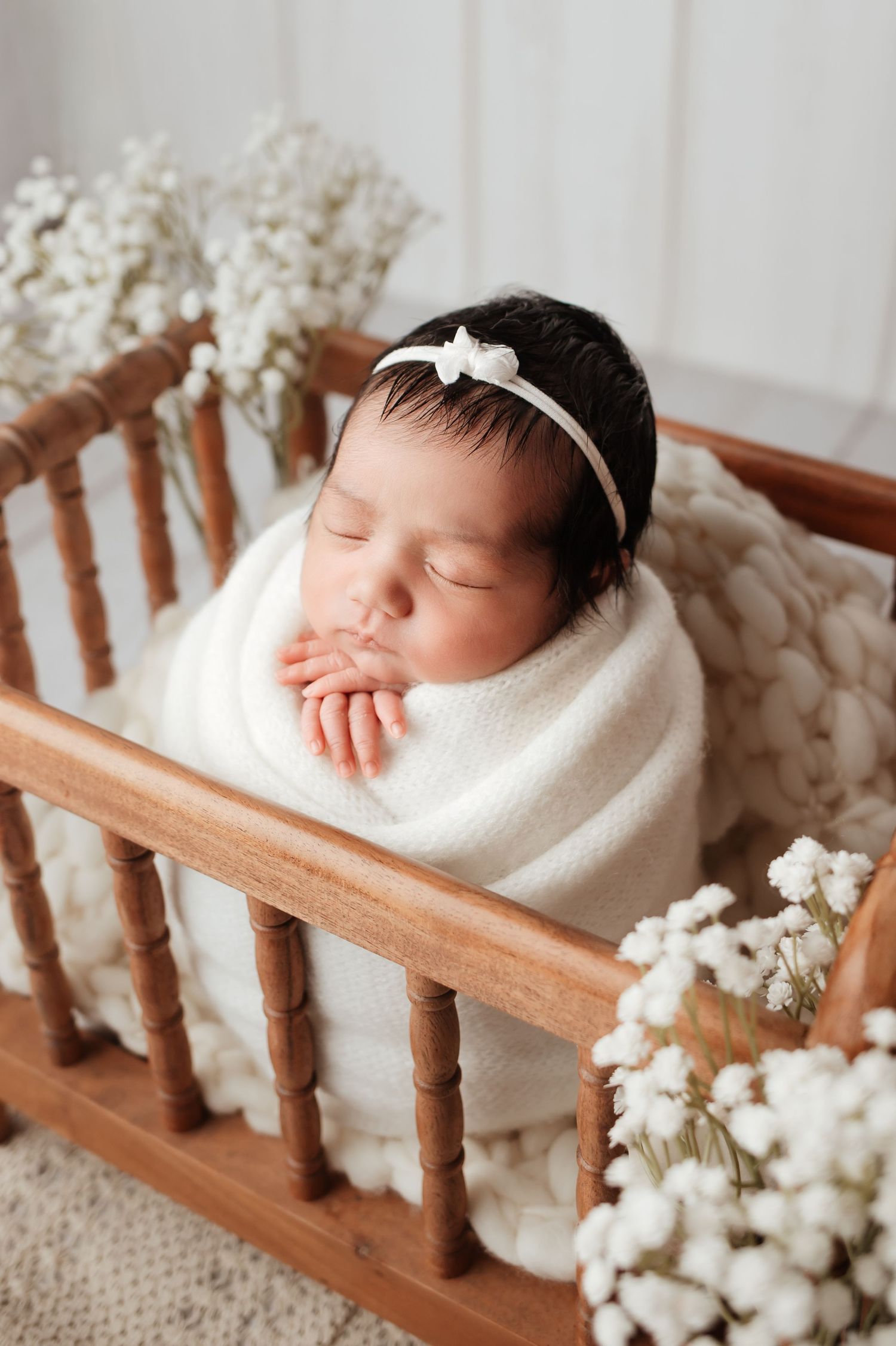 Sleeping newborn baby girl swaddled in white wrap with bow headband in wooden mini crib with white flowers.
