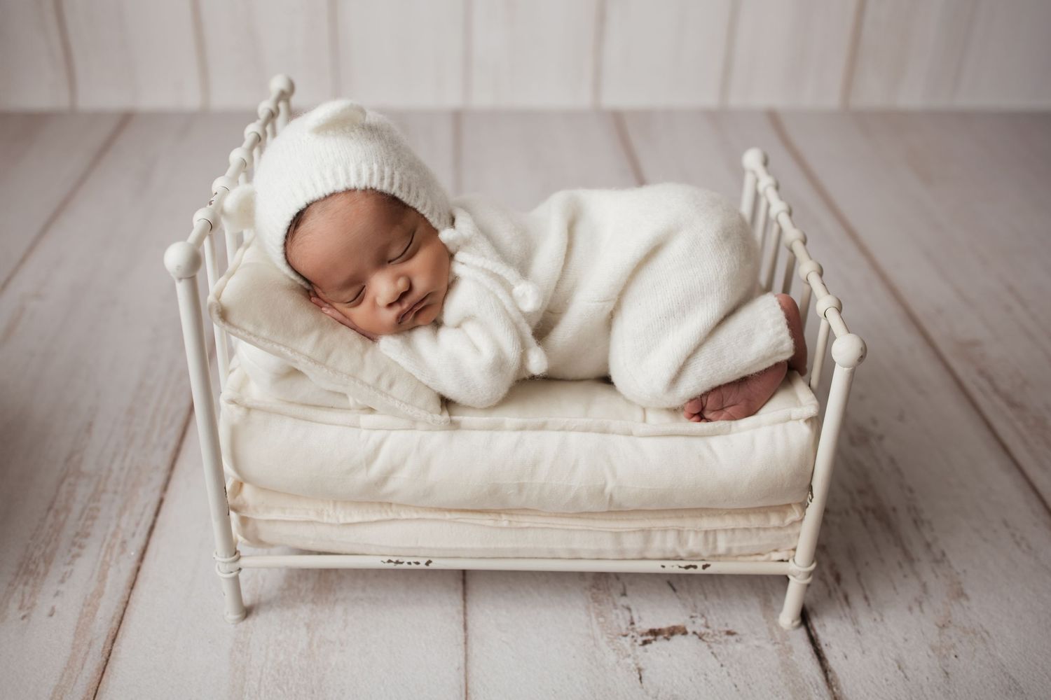 Newborn baby in white fuzzy romper and knit bonnet sleeping peacefully on small white bed.