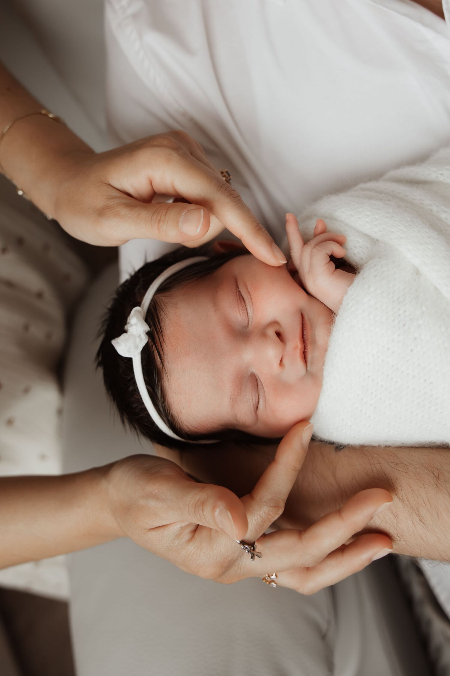 Newborn baby girl with white bow headband cradled gently in parents hands during lifestyle session.