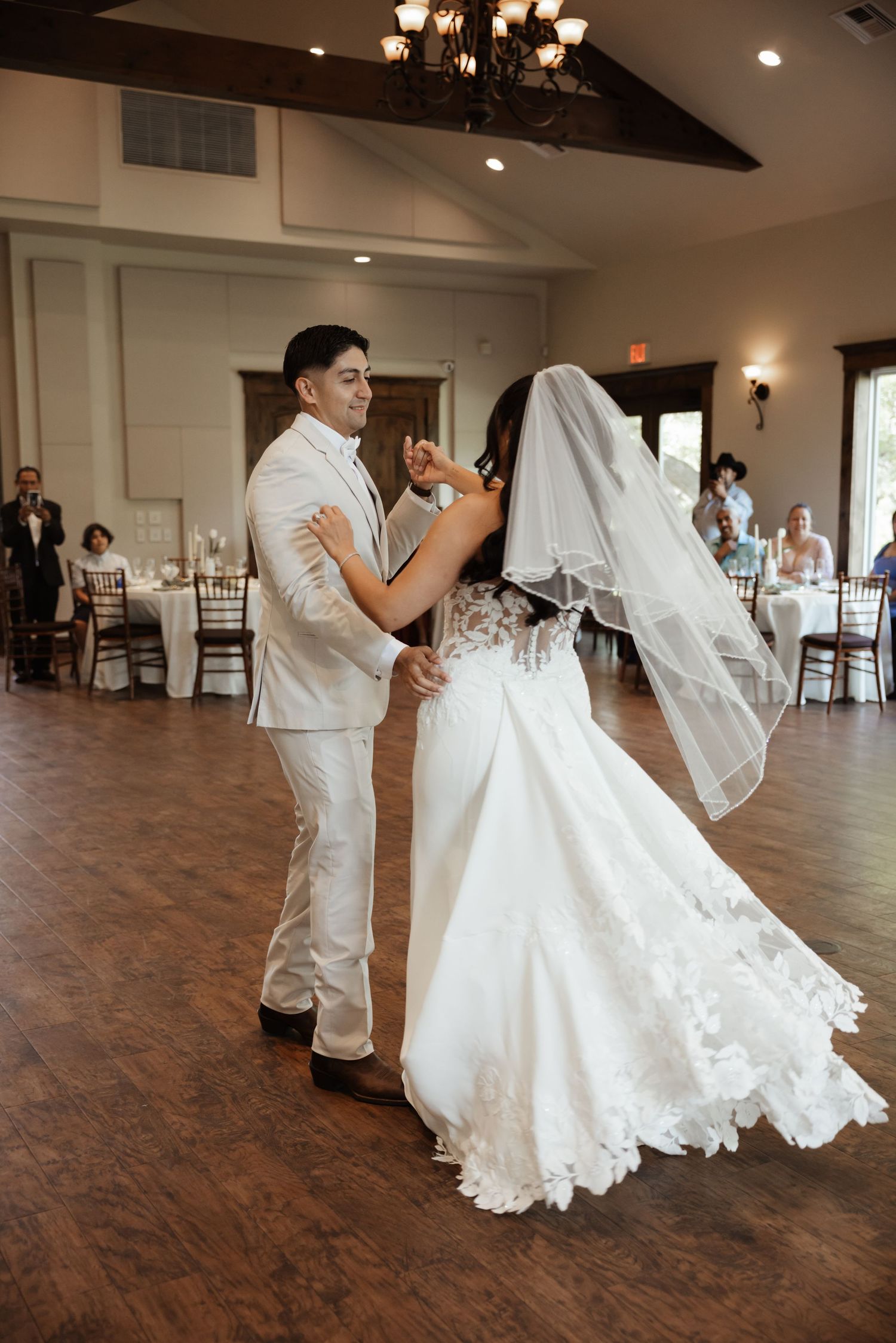 Bride and groom share their first dance in an elegant wedding reception hall with wood floors and chandelier lighting.