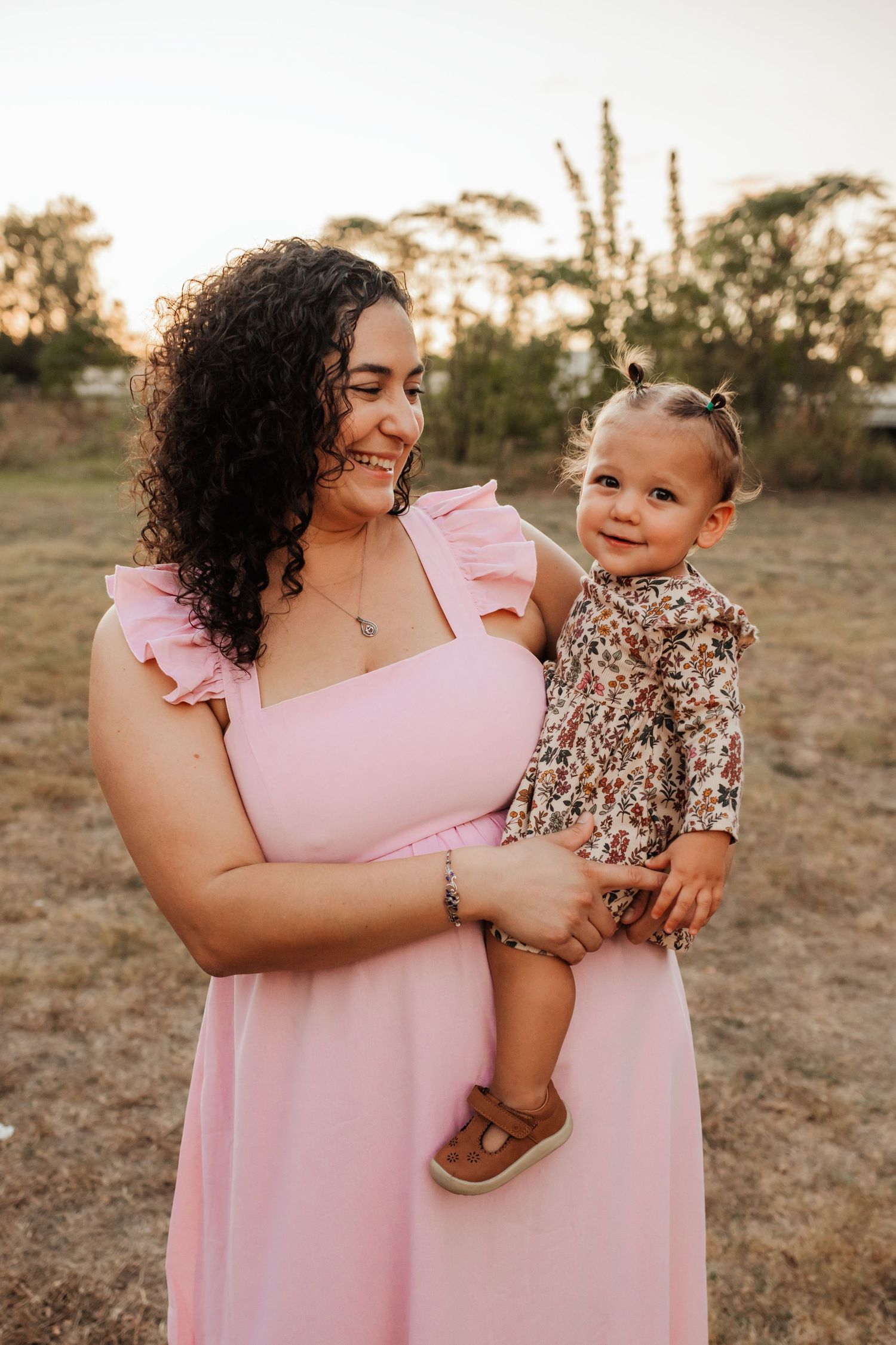 A smiling mother in a pink dress holds her happy baby girl during a warm sunset outdoor portrait.