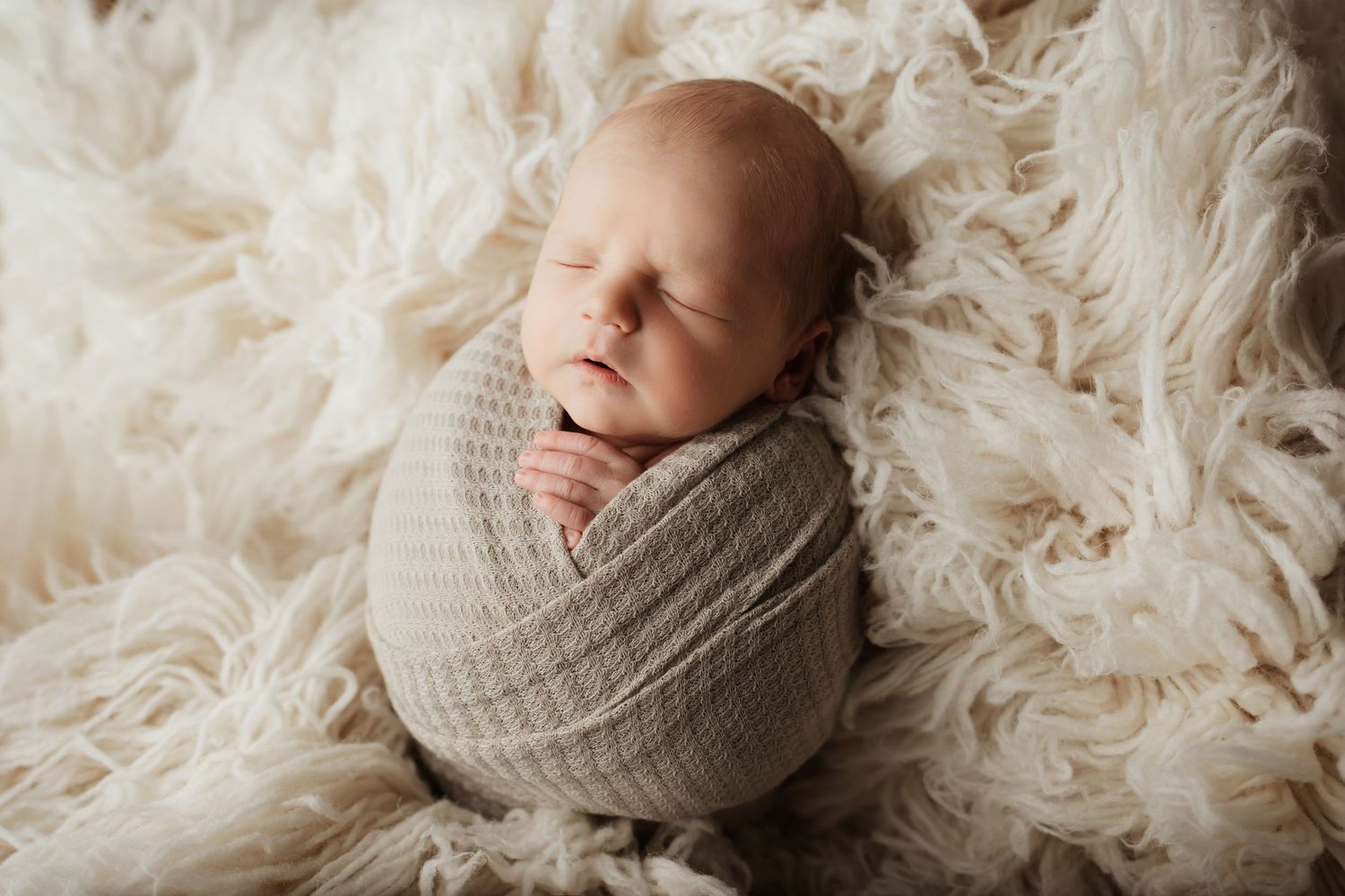 Sleeping newborn baby swaddled in a soft gray knit wrap on a fluffy cream fur blanket.