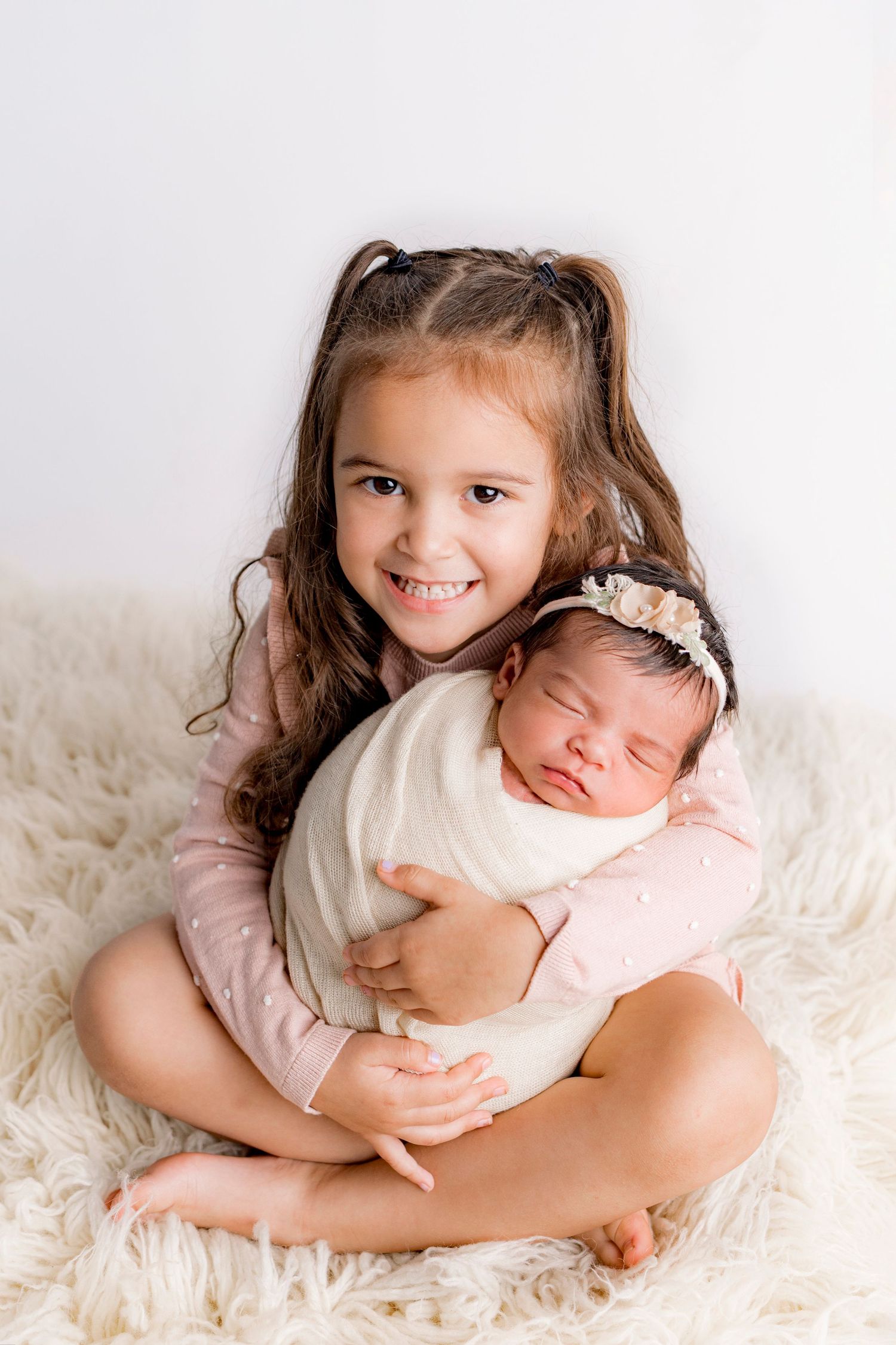 Smiling toddler girl in pink outfit holding sleeping swaddled newborn baby sister on fur rug.