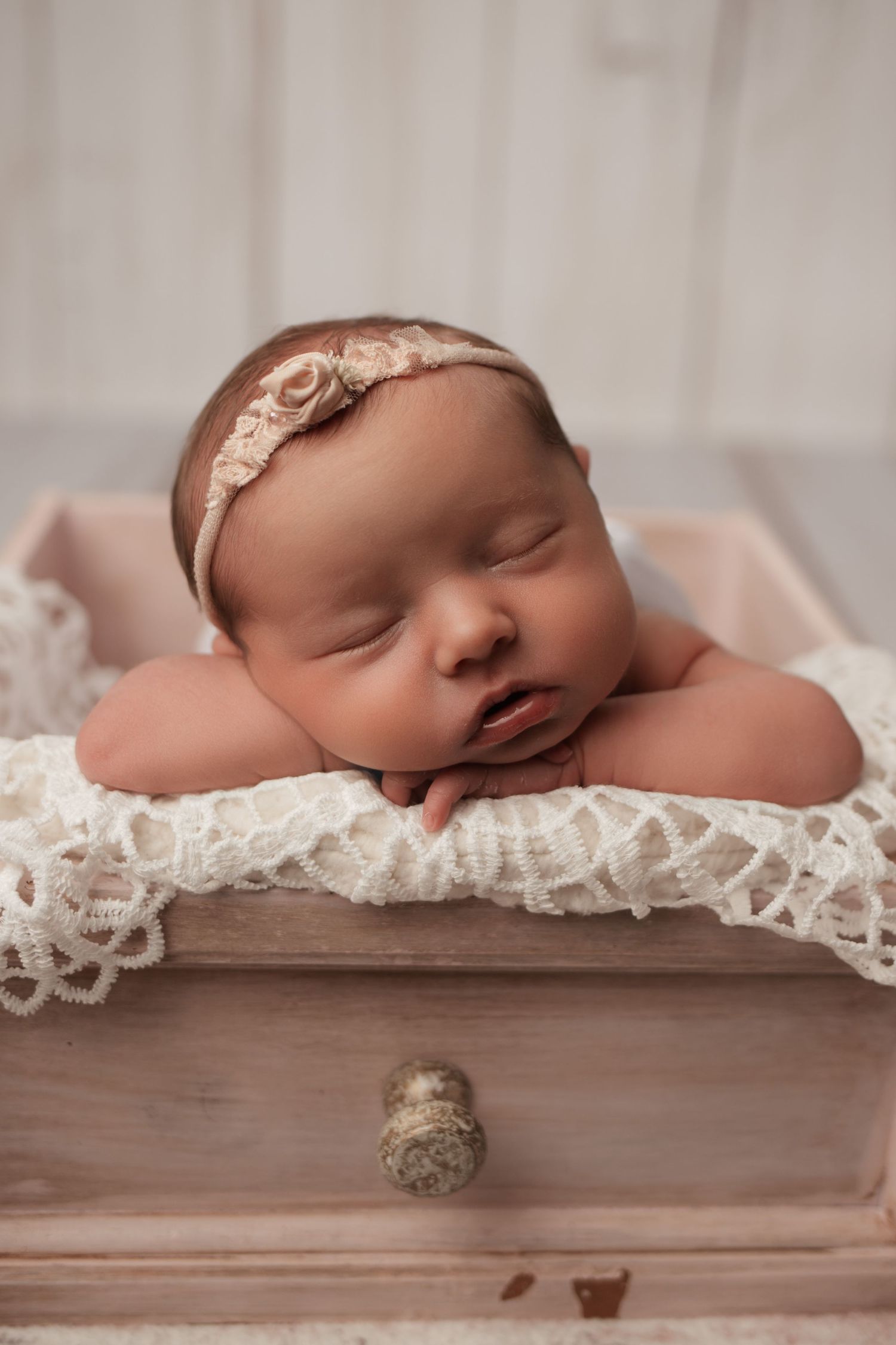 Newborn baby girl with floral headband sleeping with chin resting on lace in drawer.