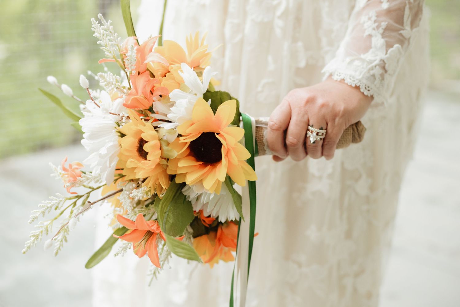 Bride in lace gown holding a sunflower and peach lily wedding bouquet with white blooms, wearing a diamond ring.