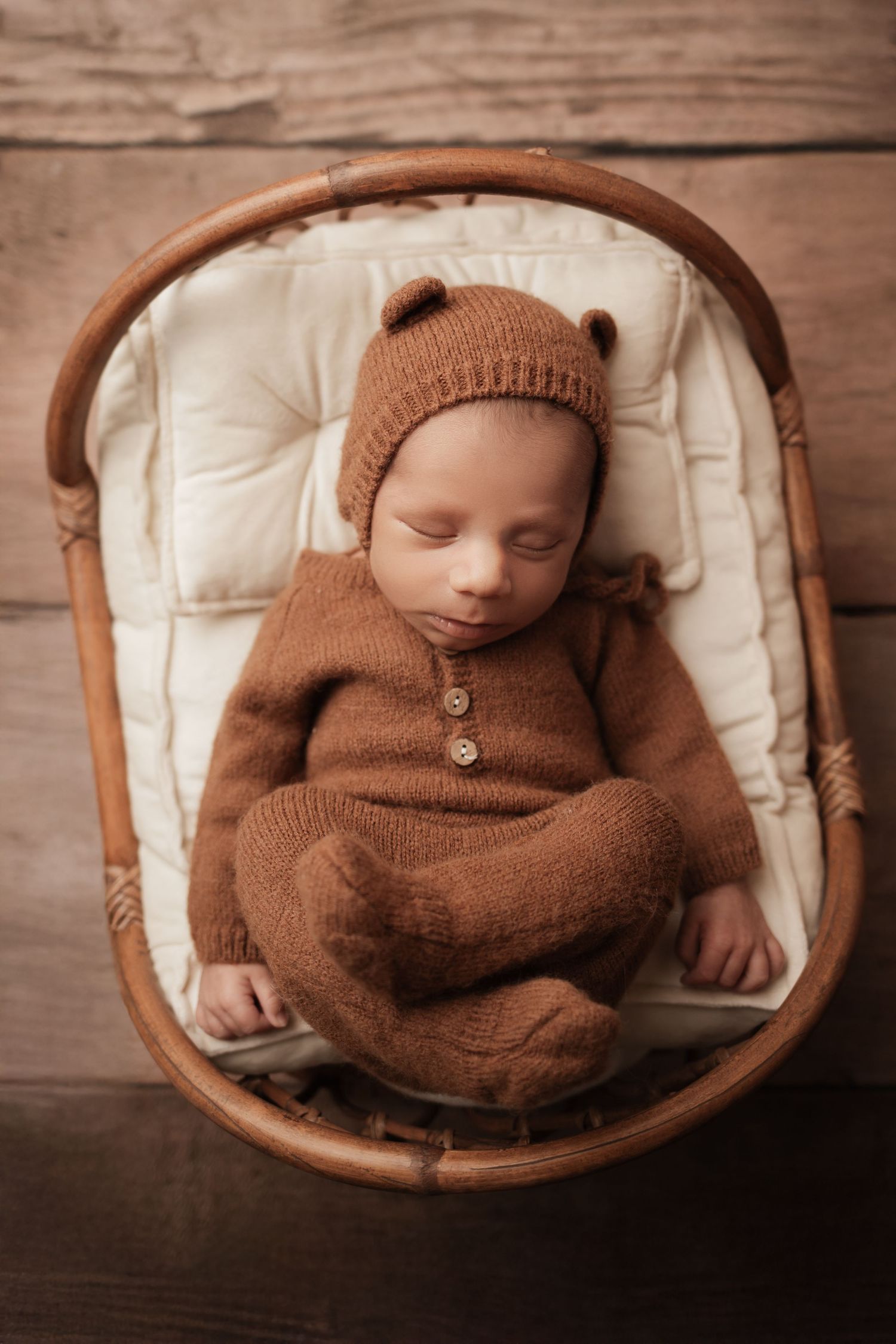 Newborn baby in brown knit bear outfit and bonnet sleeping in wicker bassinet on wood floor.