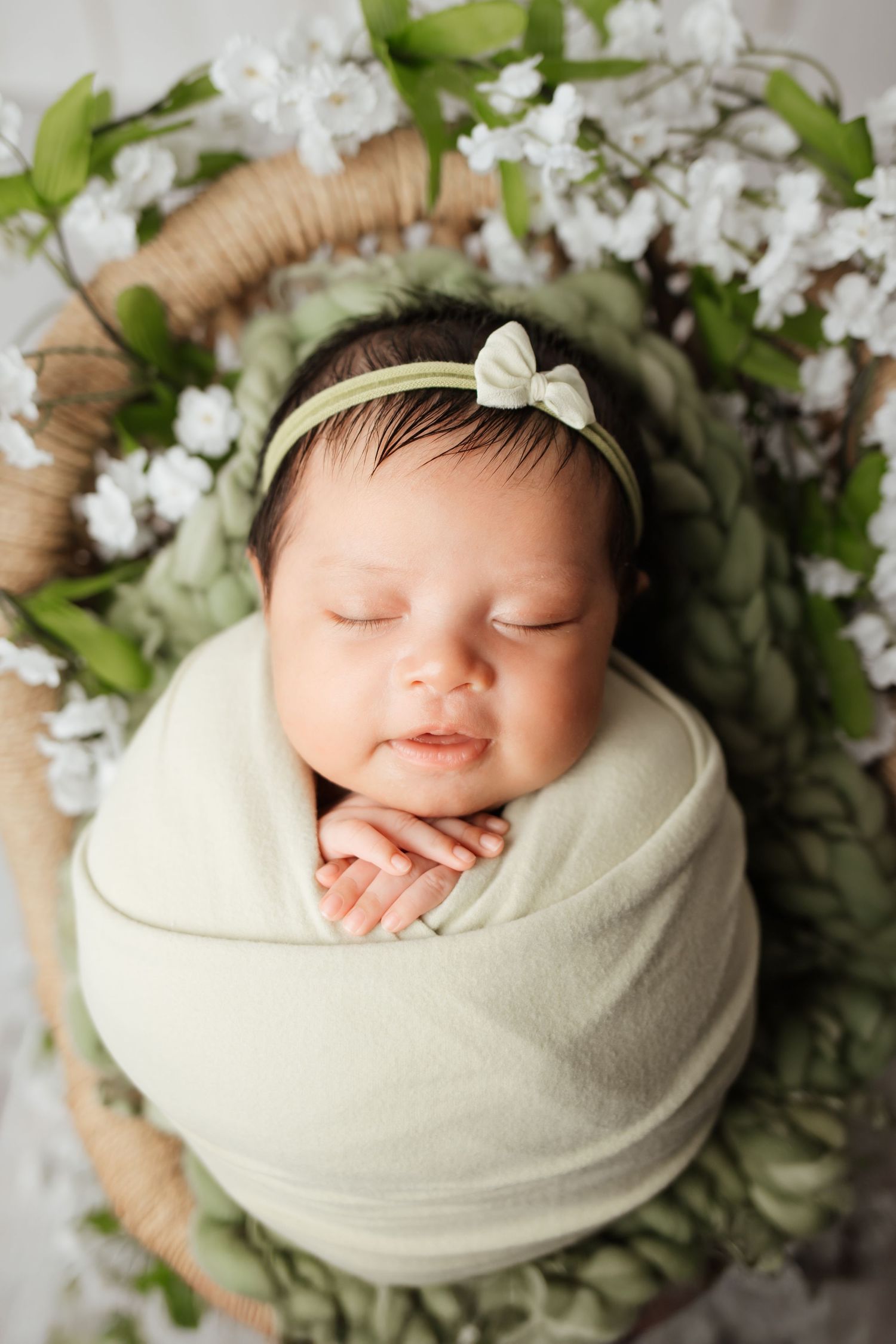 Sleeping newborn baby girl swaddled in green wrap with a bow headband in a floral basket.