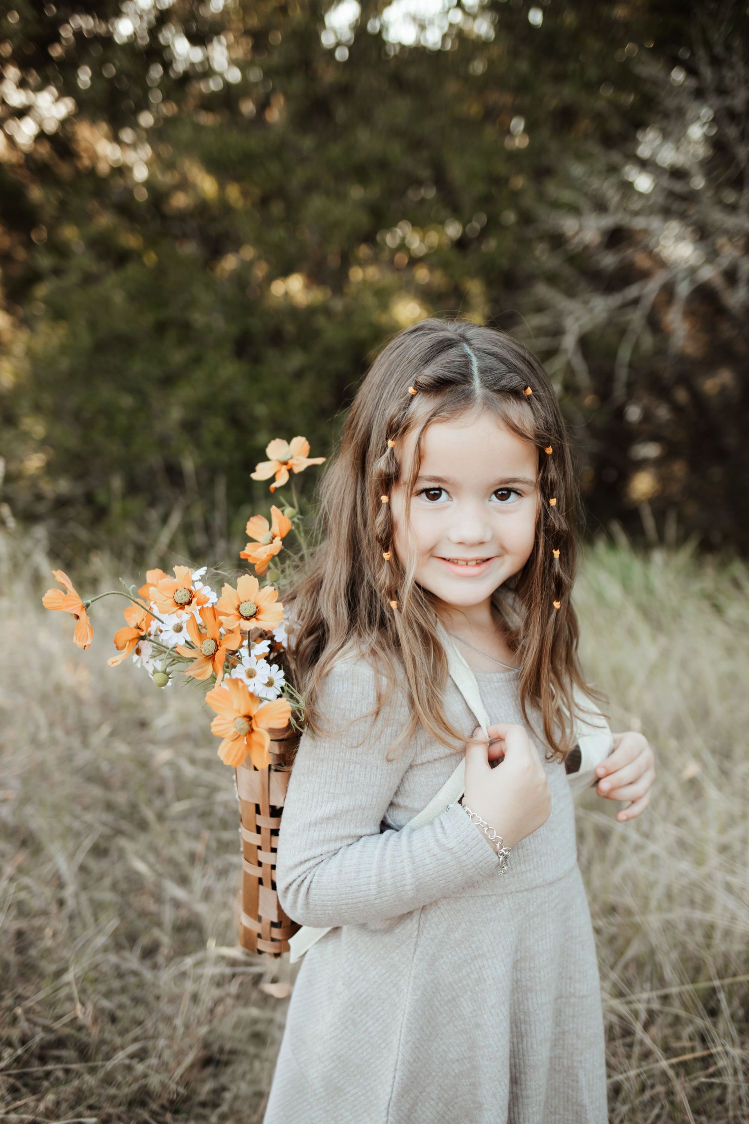 Smiling young girl in a gray dress holding a basket of orange wildflowers in a sunlit field.