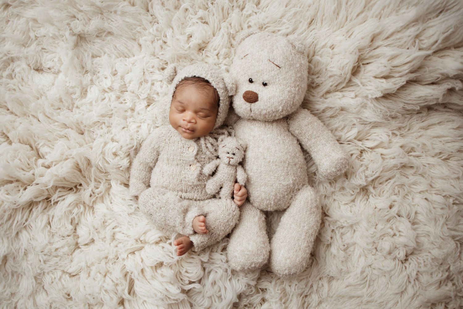 A sleeping newborn in a cream teddy bear outfit cuddles next to a matching stuffed bear on fur.