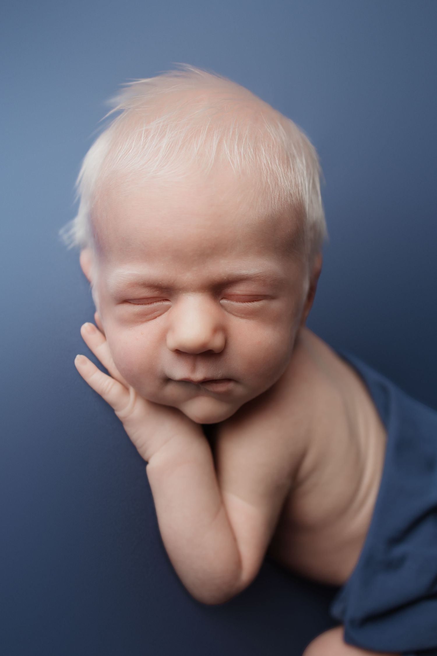 Close-up of a blonde newborn baby resting chin on hands while sleeping on a blue background.