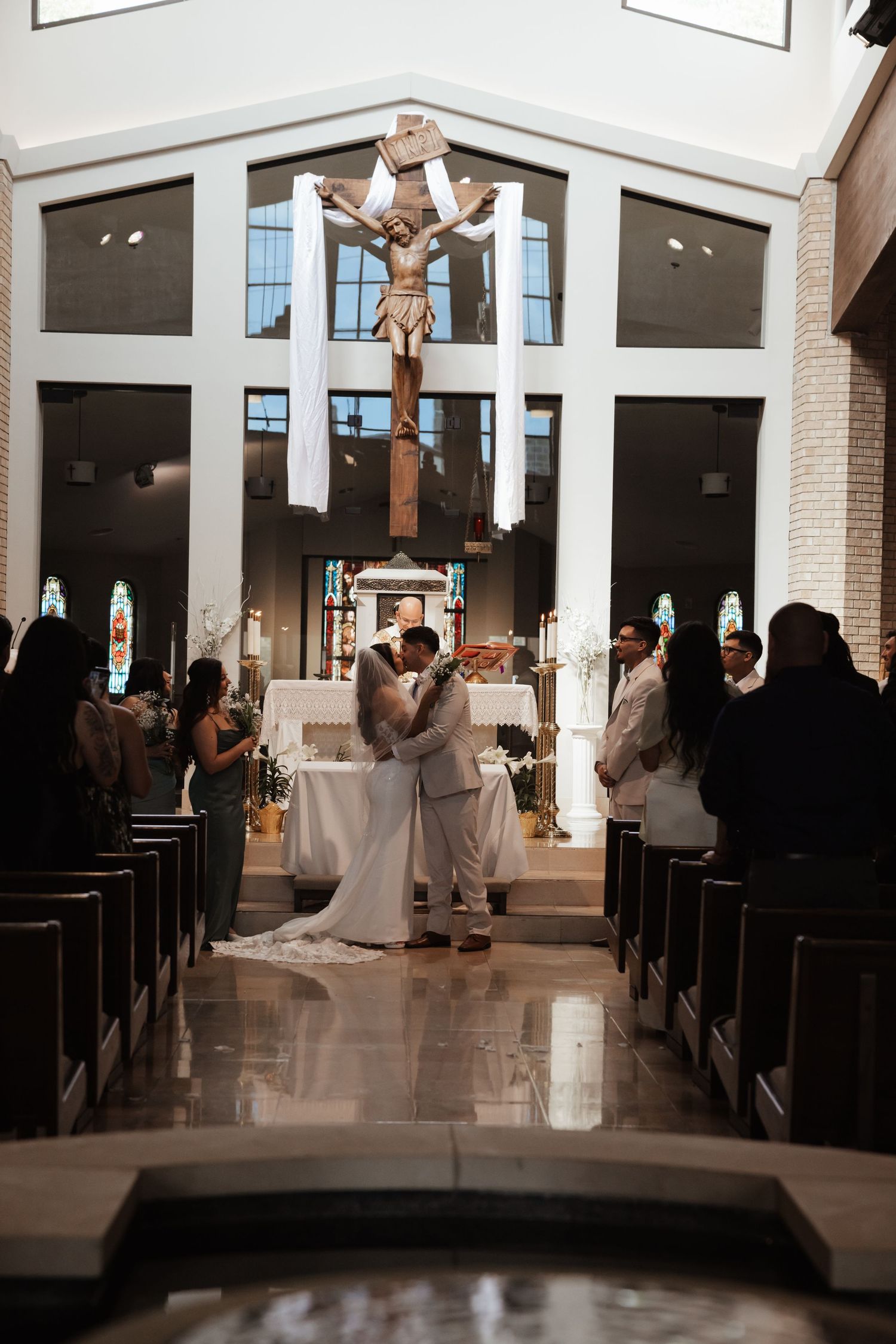 Bride and groom share first kiss at Catholic church altar with crucifix backdrop during wedding ceremony.