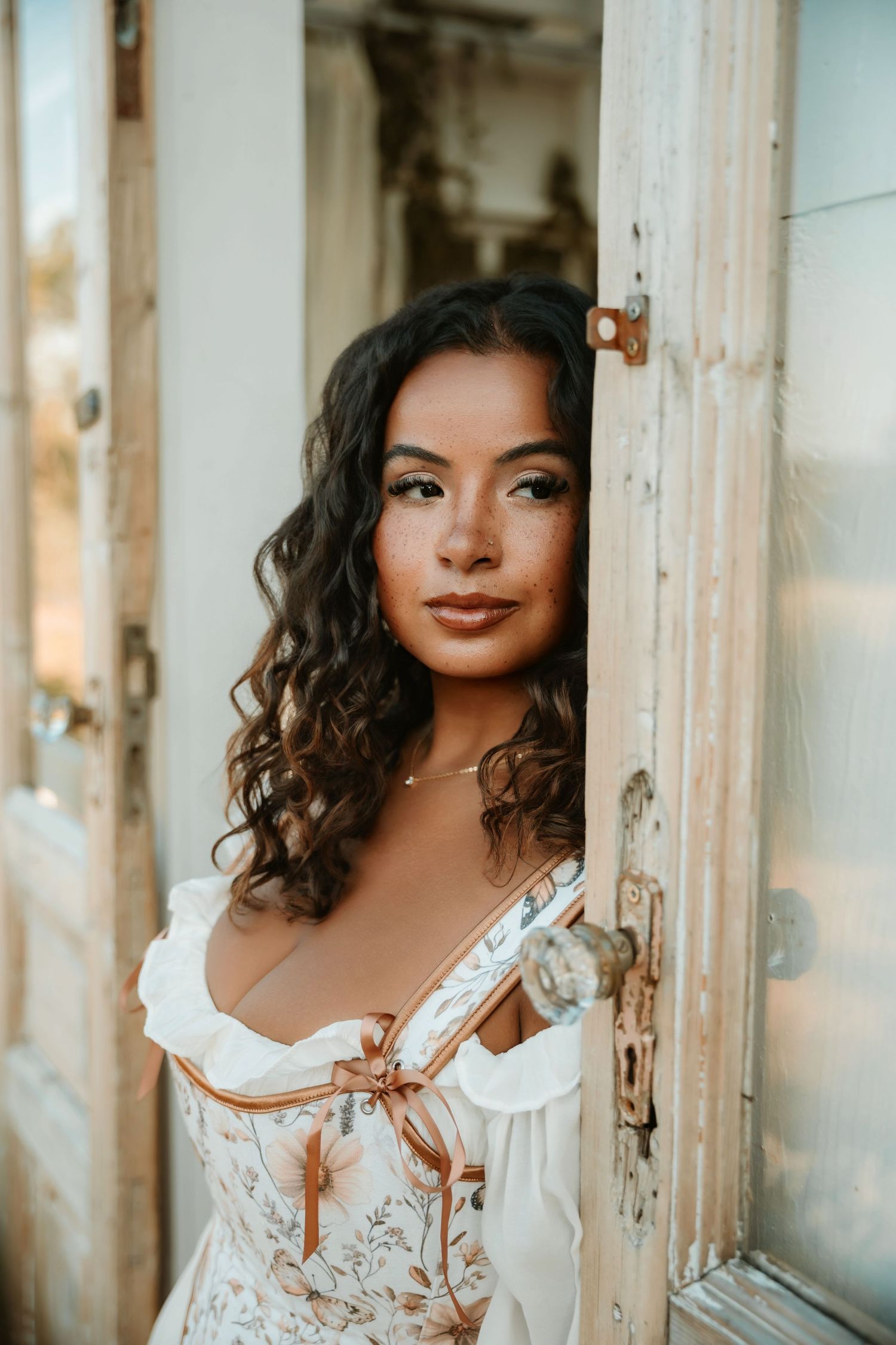 Young woman with curly dark hair in a white floral corset dress poses in a rustic doorway.