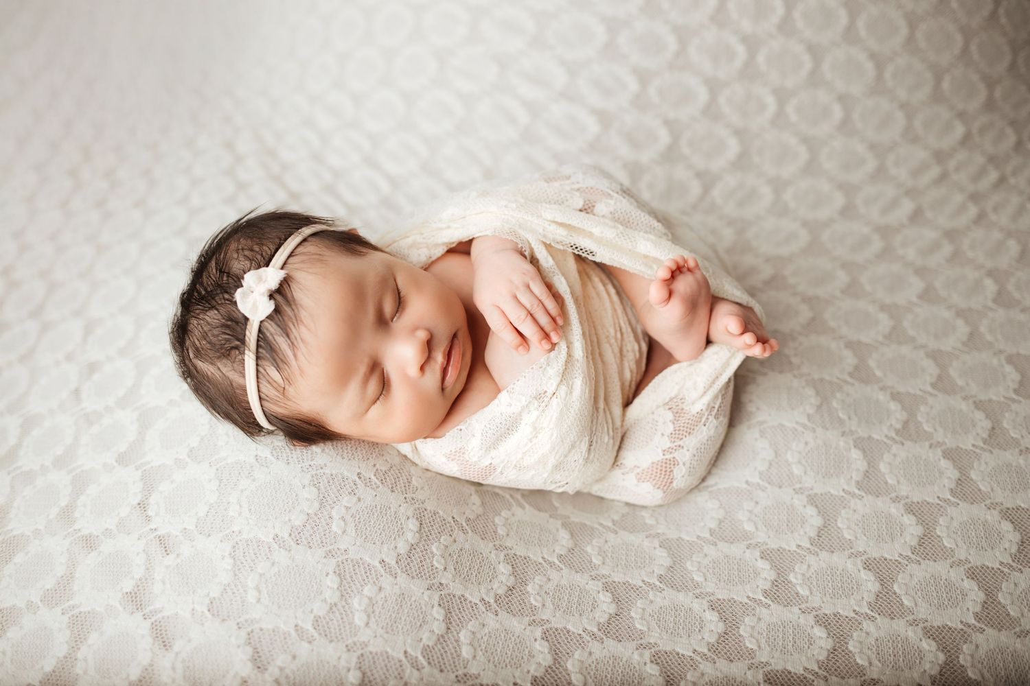Peaceful newborn baby girl swaddled in white lace wrap, wearing a bow headband, sleeping on a lace blanket.