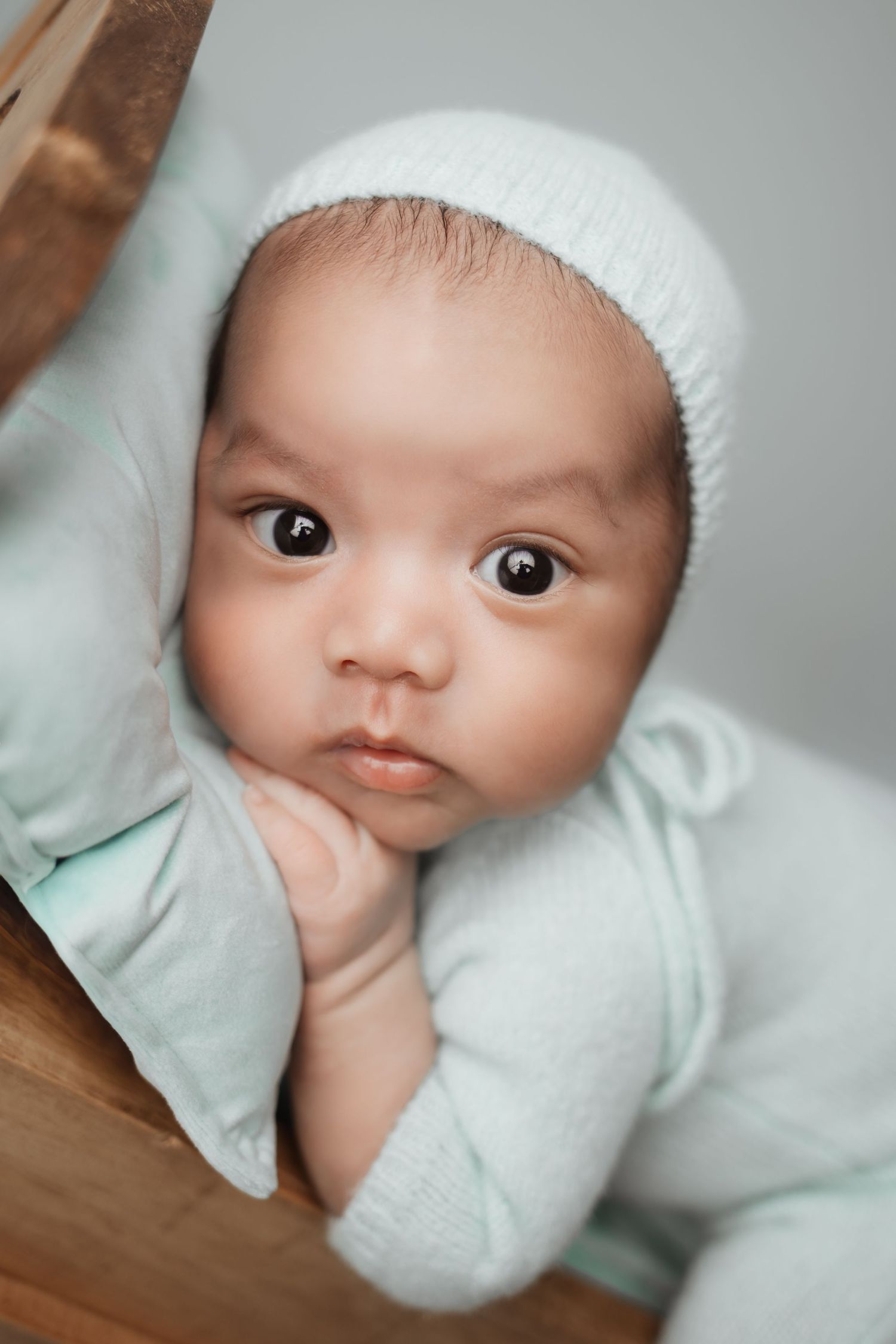 Alert newborn baby with big dark eyes wearing mint green bonnet and outfit in wooden crib.