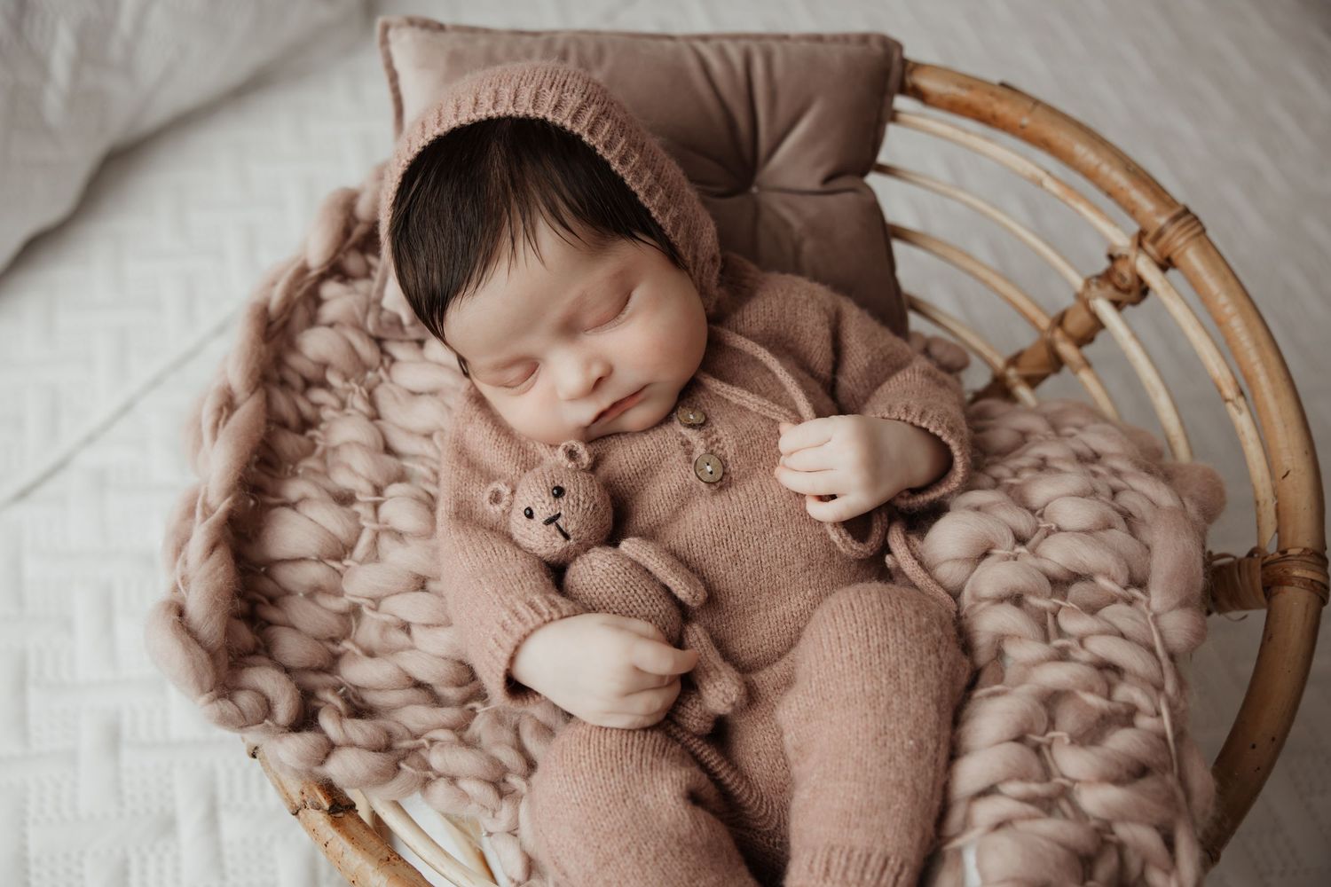 Newborn baby sleeping in a wicker chair wearing a brown knit bonnet and onesie holding a teddy bear.