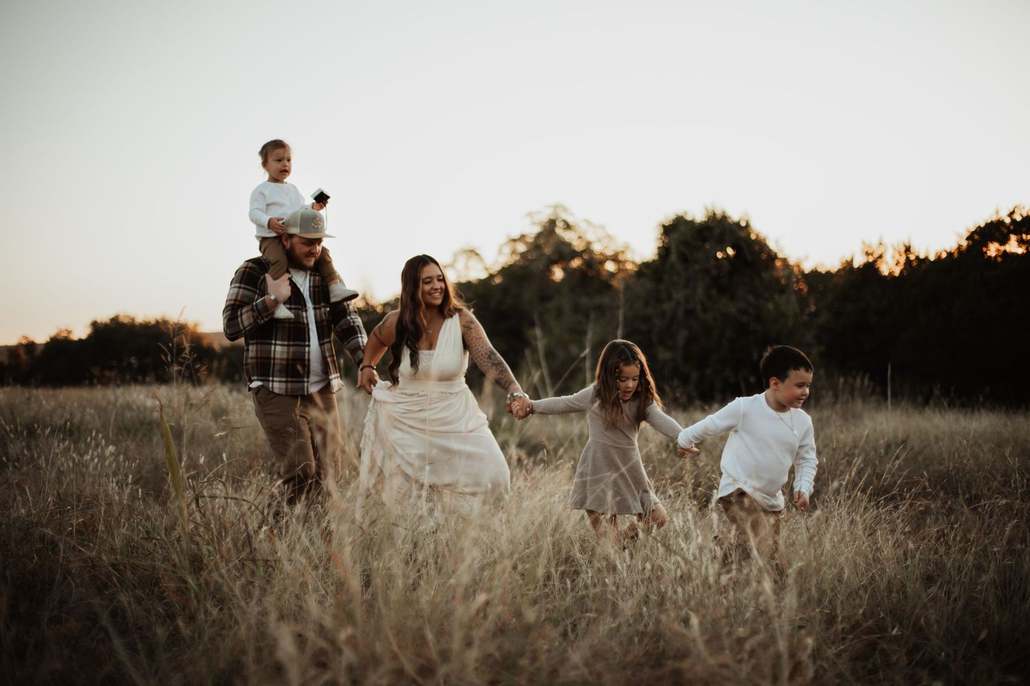 A joyful family of five walks hand-in-hand through a golden field at sunset in a candid outdoor portrait.
