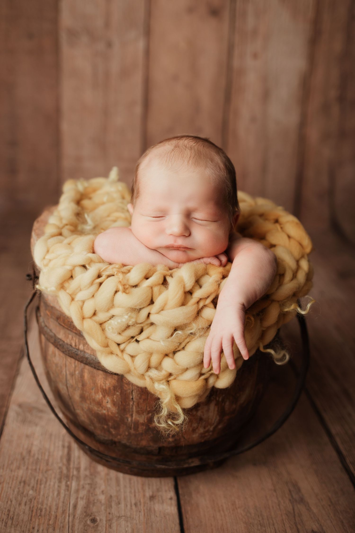 Adorable sleeping newborn baby posed in a rustic wooden bucket with chunky yellow knit blanket.