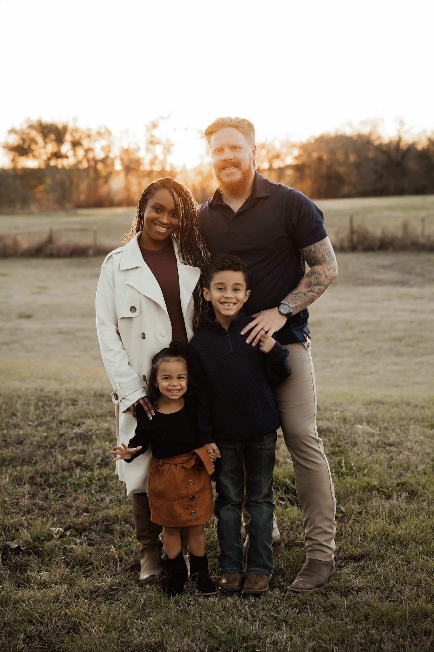 A happy interracial family of four poses together in a field during golden hour sunset.