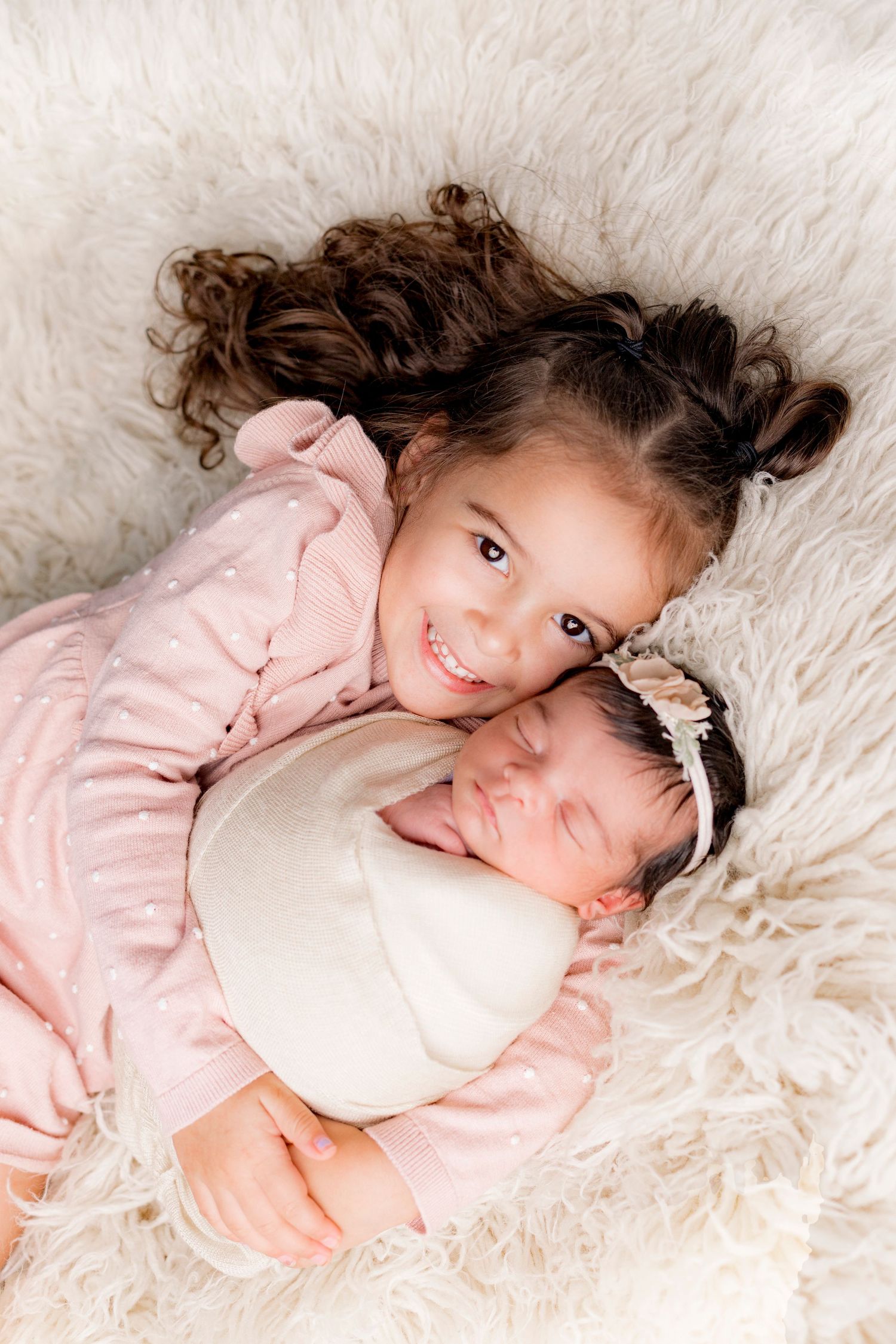 Happy toddler girl in pink dress lying with sleeping swaddled newborn baby sister on fluffy rug.