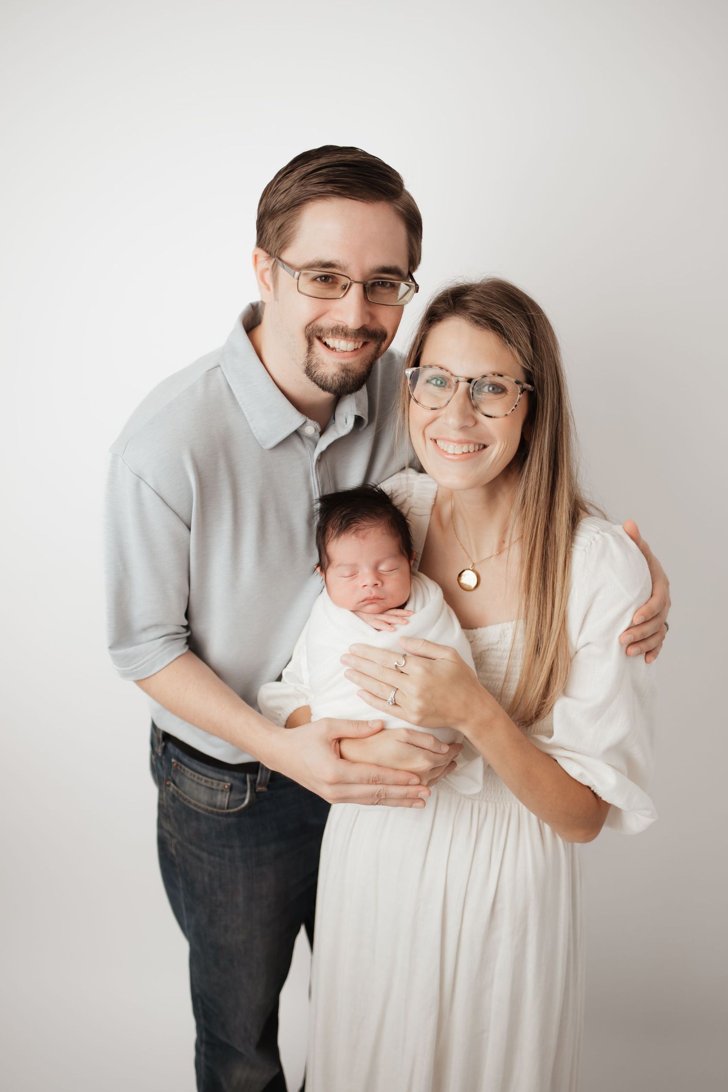 Happy parents holding their sleeping newborn baby wrapped in white during studio portrait session.