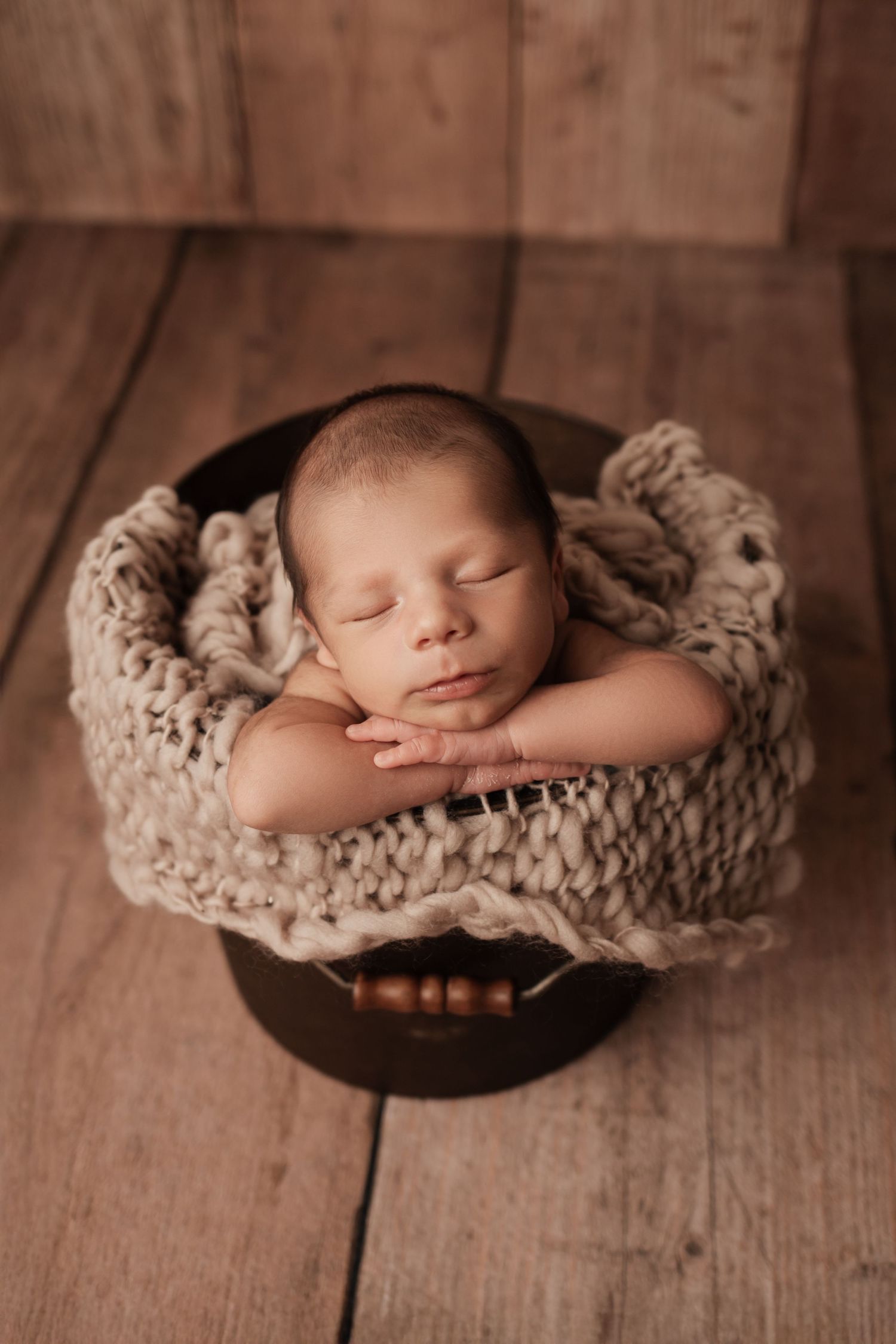 Sleeping newborn baby posed in rustic wooden bowl with chunky knit blanket on wood floor.