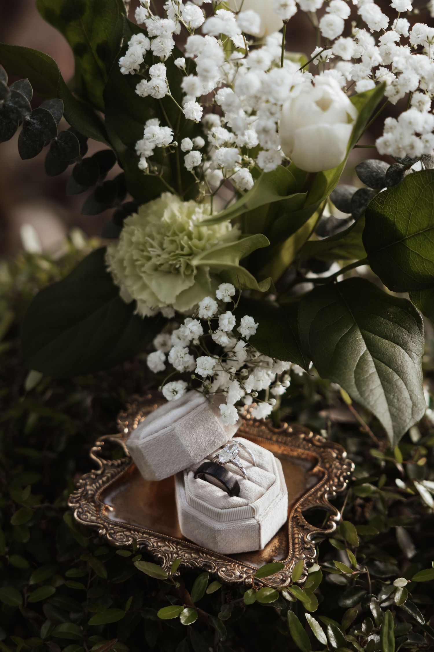 Wedding rings in a white velvet box on a vintage tray surrounded by a bridal bouquet of white flowers and greenery.