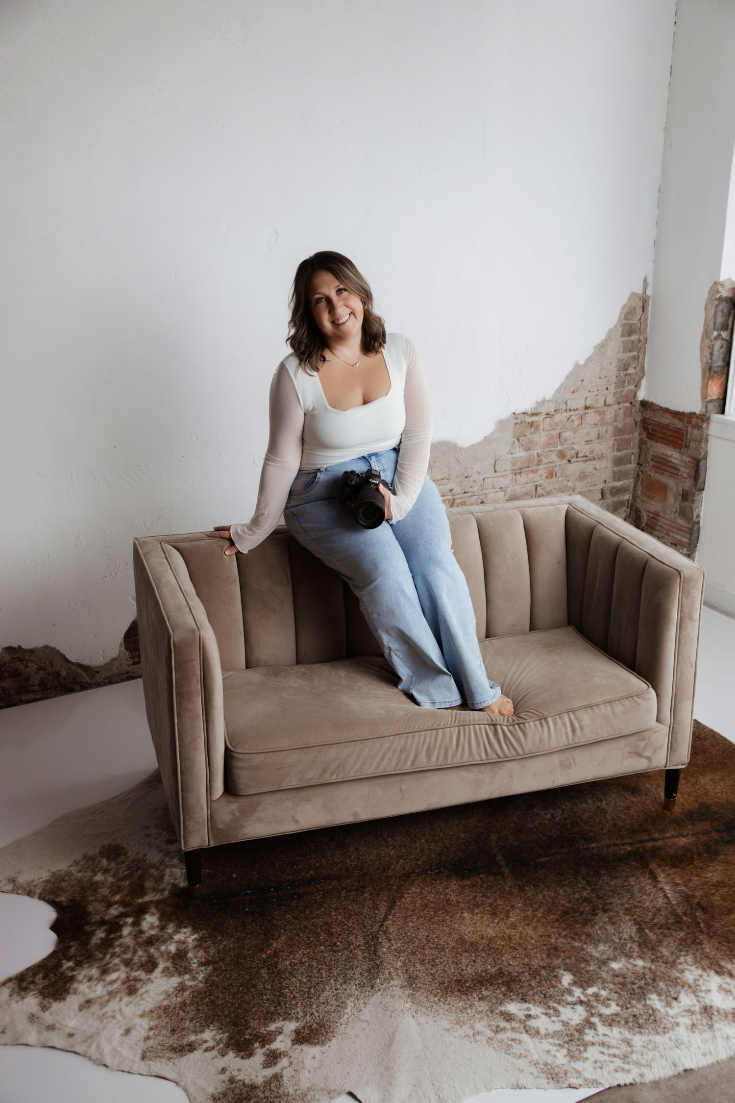 Woman in white top and jeans sitting on a beige velvet sofa in a rustic studio with exposed brick wall.