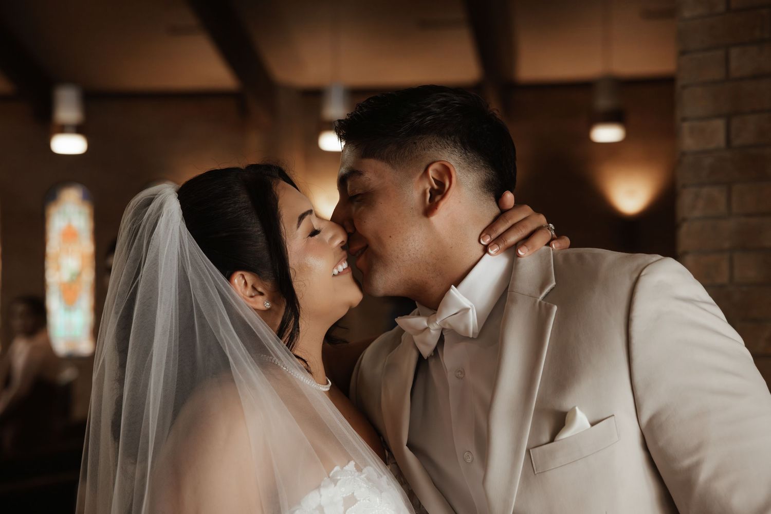 Groom in beige suit kisses bride in white dress and veil during romantic wedding ceremony in church.