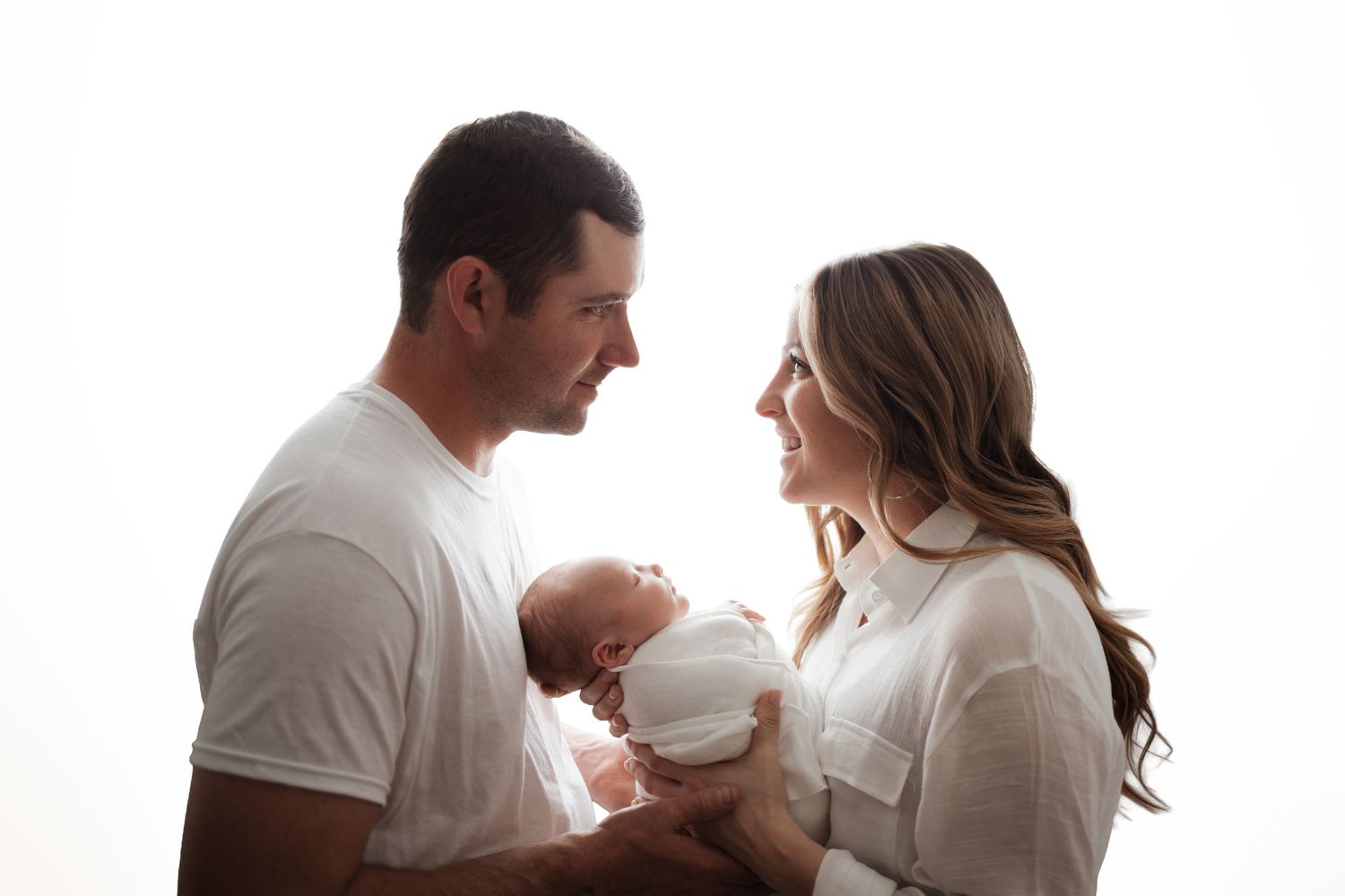 A happy couple in white clothing smiles at each other while gently holding their swaddled newborn.