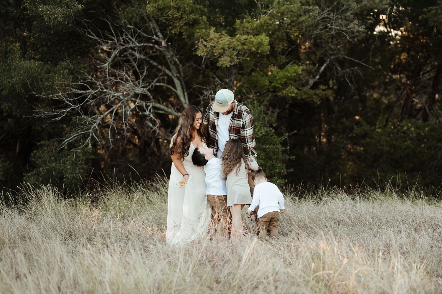 A family shares a tender moment together in a golden grass field surrounded by trees at dusk.