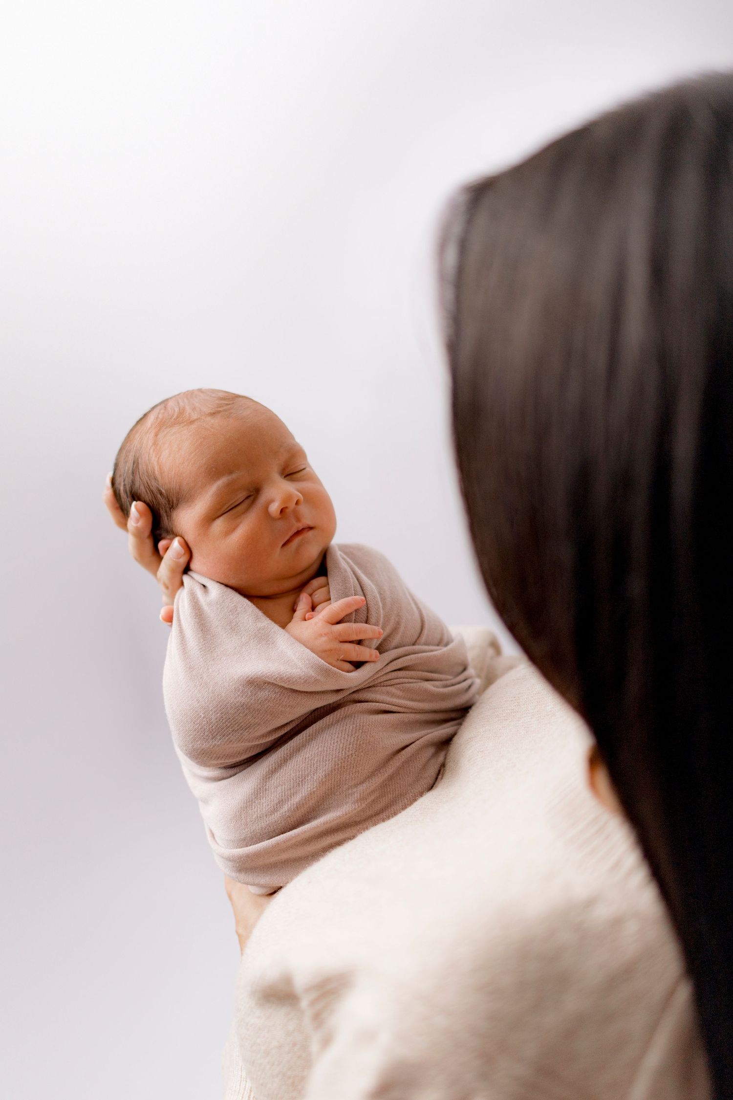 Mother with dark hair holding sleeping newborn baby wrapped in beige swaddle against white background.