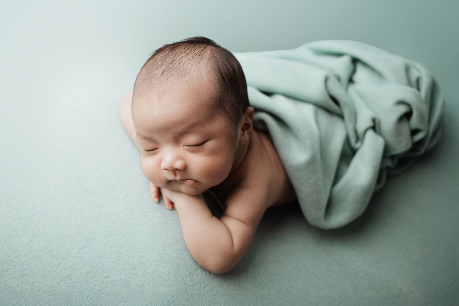 Newborn baby sleeping on teal surface partially wrapped in matching green blanket, hands tucked under chin.