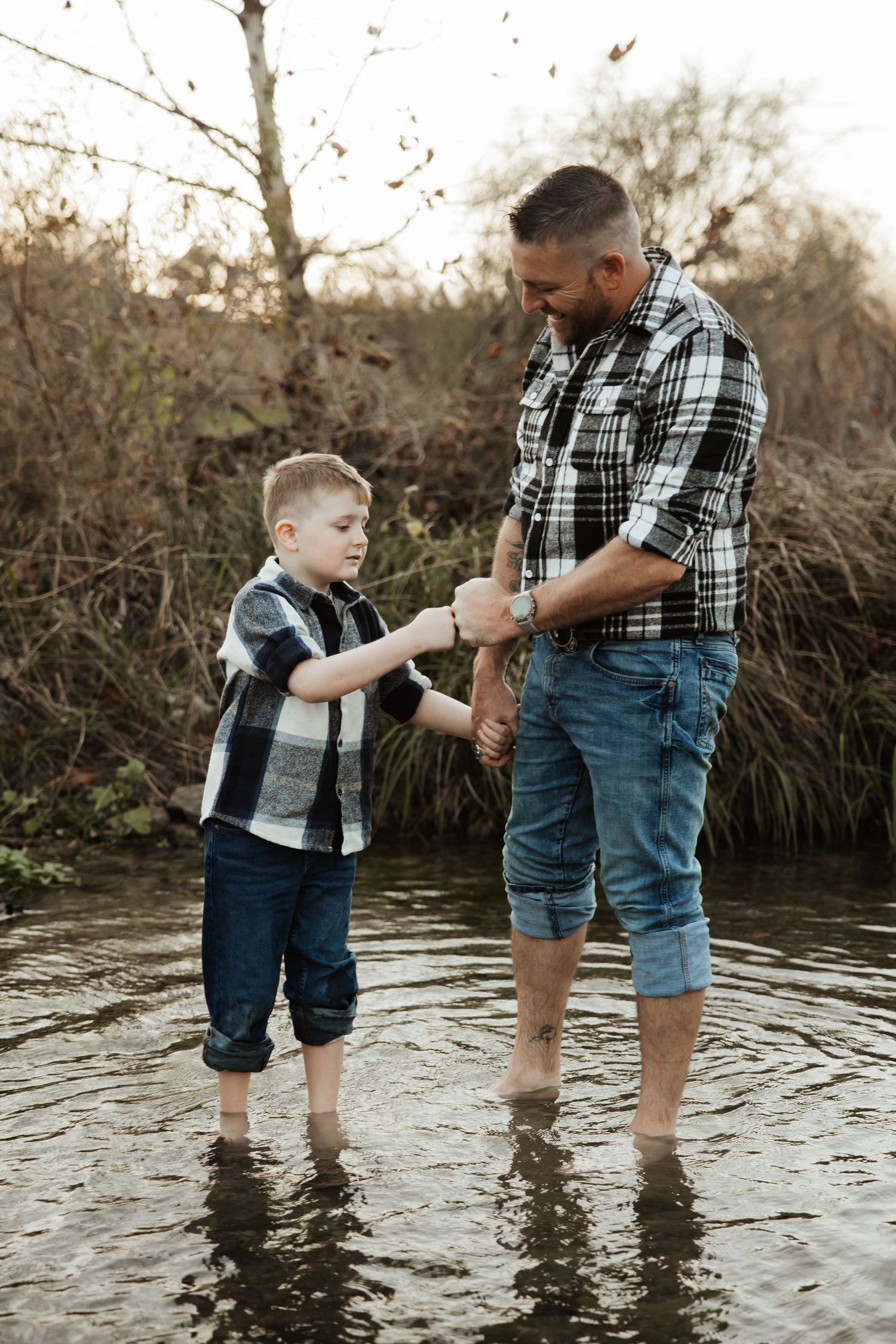A father and son in matching plaid shirts stand holding hands in a shallow creek.