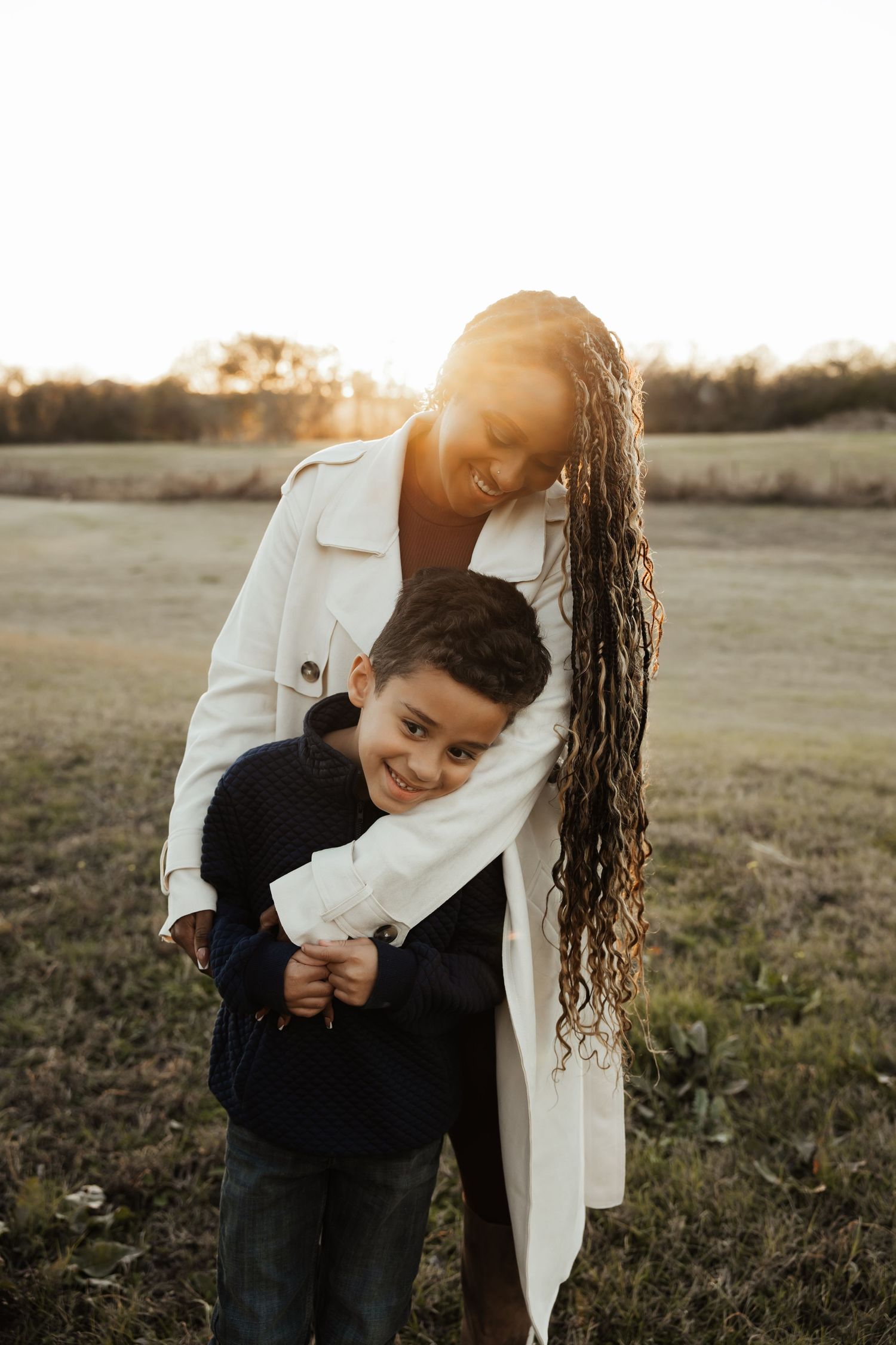 A joyful Black mother with long braids hugs her young son outdoors at golden hour.