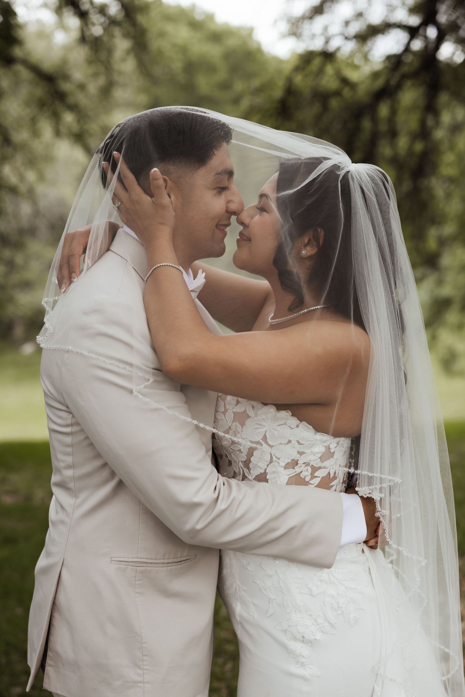 Bride and groom sharing a romantic moment under a wedding veil outdoors, surrounded by lush green trees.