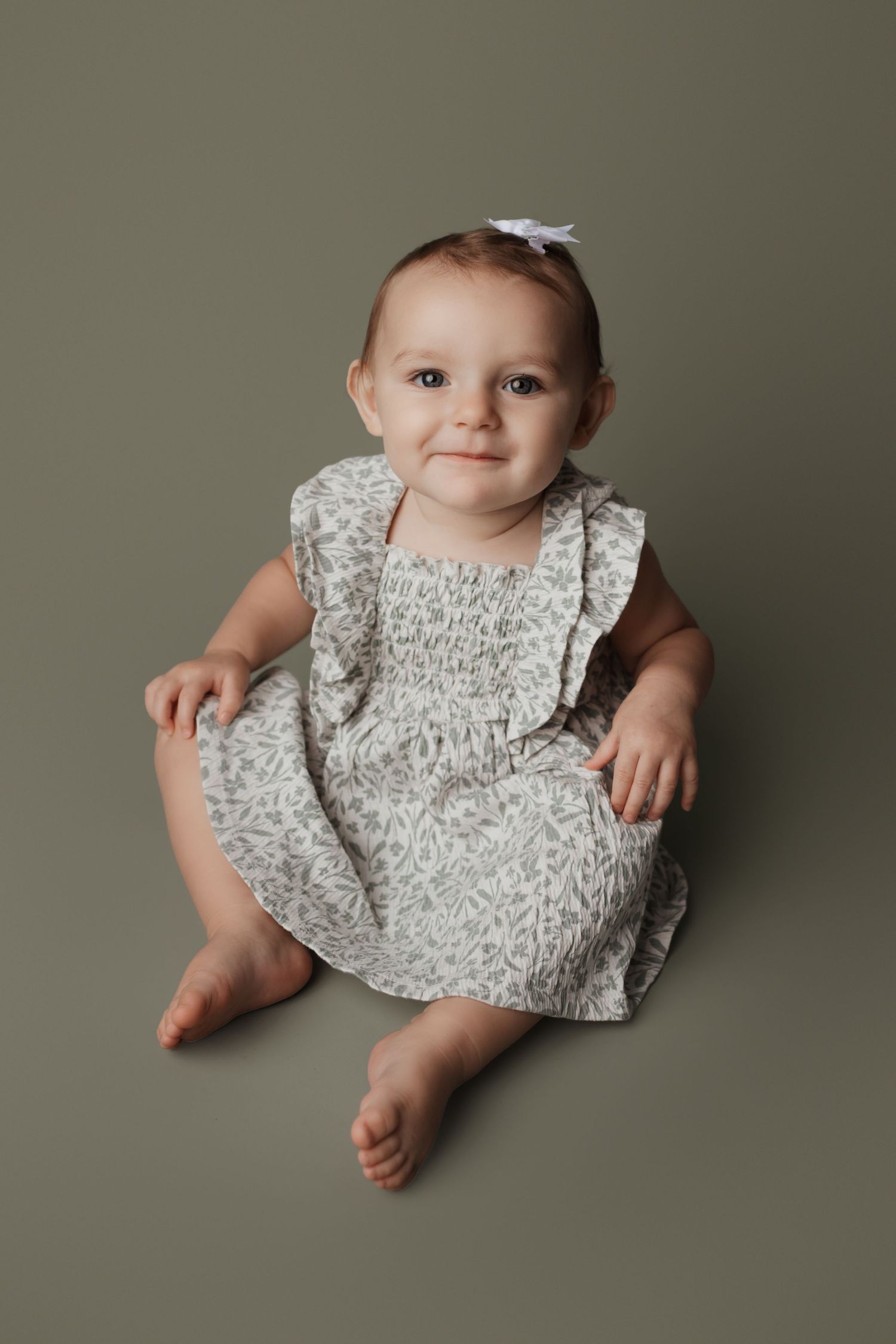 Adorable baby girl in floral dress sitting on olive green backdrop during milestone photo session.