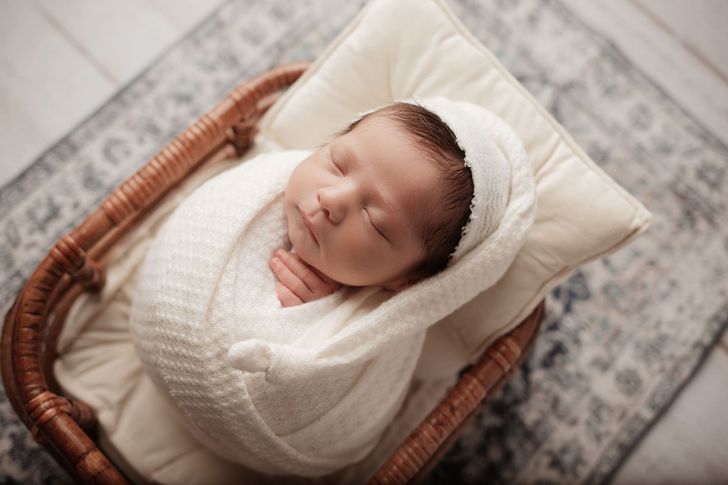 Newborn baby swaddled in white knit wrap sleeping in a wicker basket with pillow.