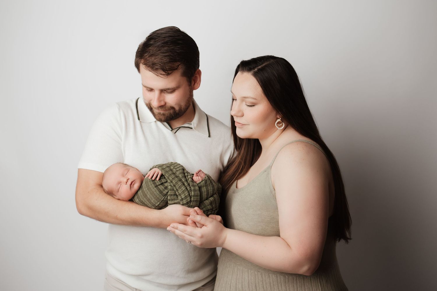 New parents lovingly gazing at their sleeping newborn baby wrapped in green against white backdrop.