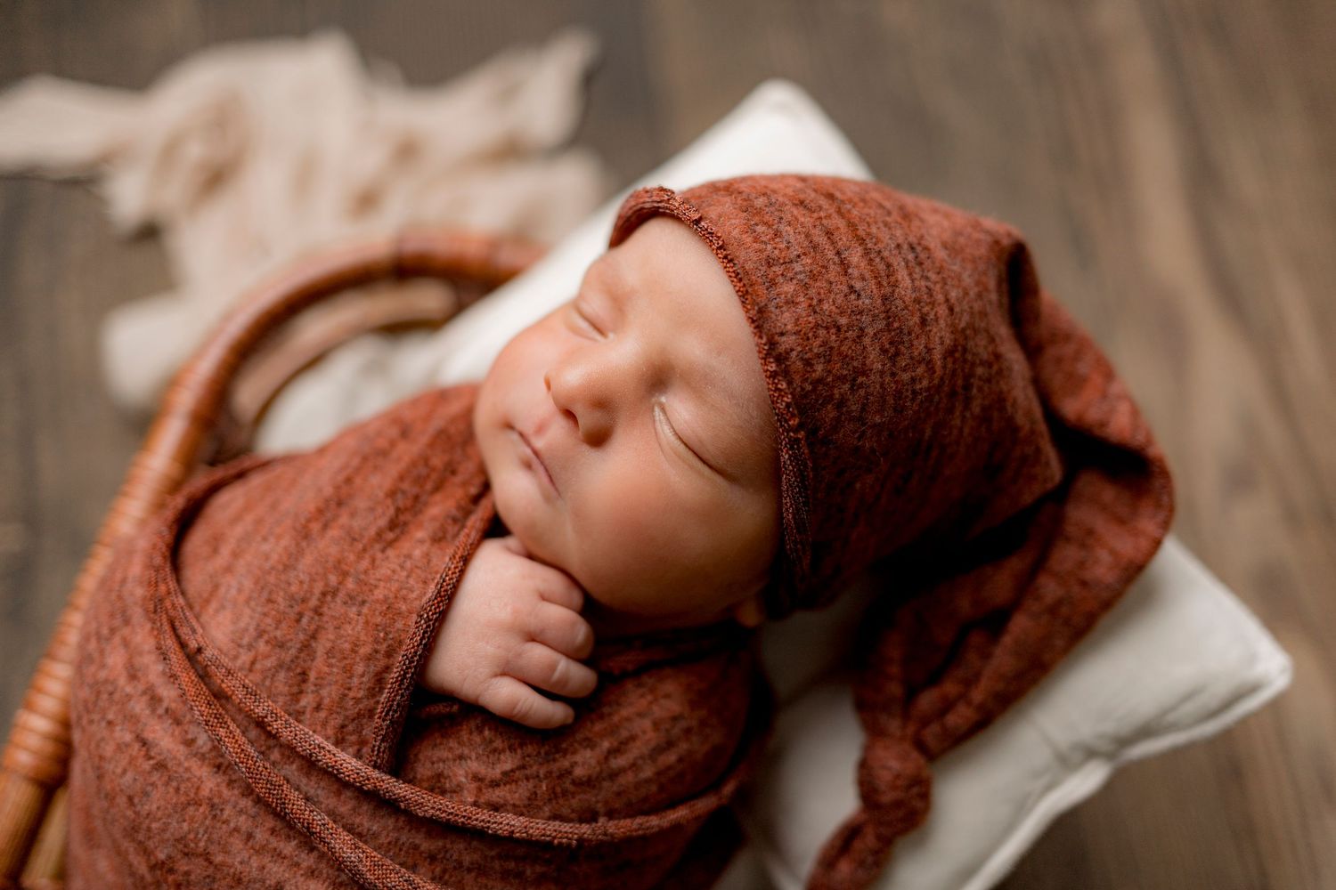 Sleeping newborn wrapped in rust orange knit swaddle and matching bonnet in wicker basket.